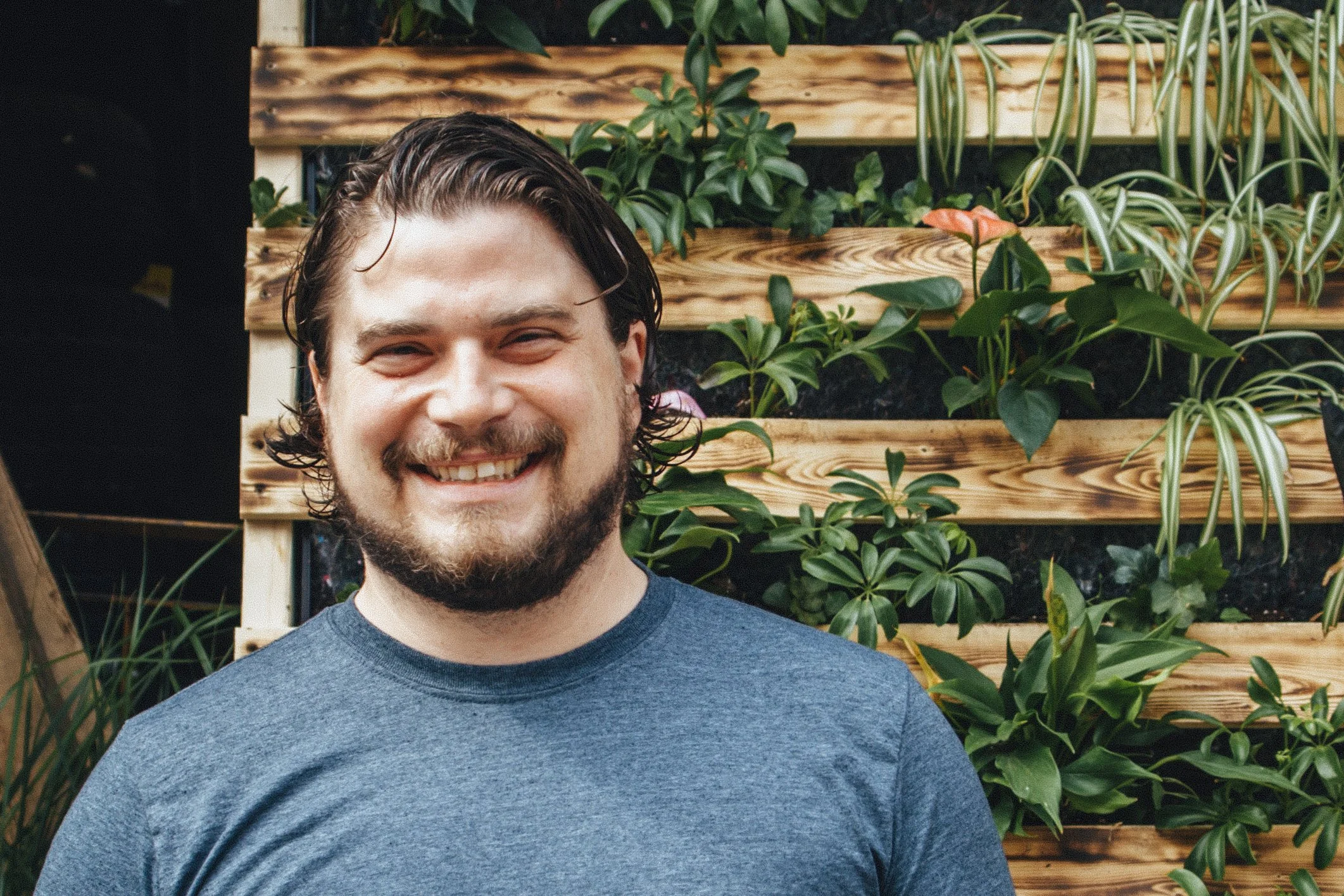 Jeune homme souriant portant un t-shirt gris foncé, derrière un mur végétal en bois avec des plantes