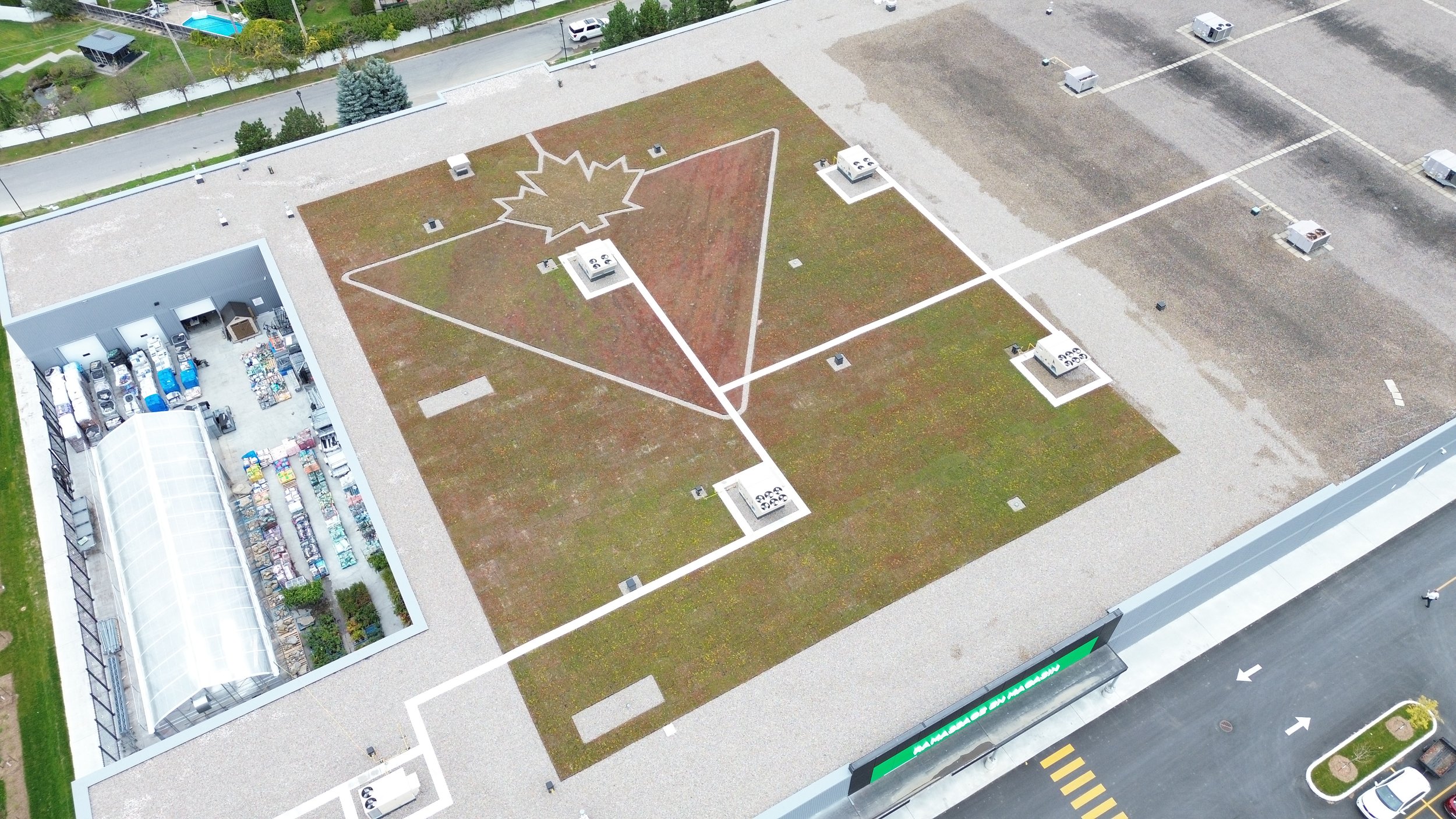 Toit plat d'un bâtiment commercial avec un espace vert, lignes blanches formant un dessin de feuille d'érable, et plusieurs unités de climatisation. Vue aérienne de la ville.