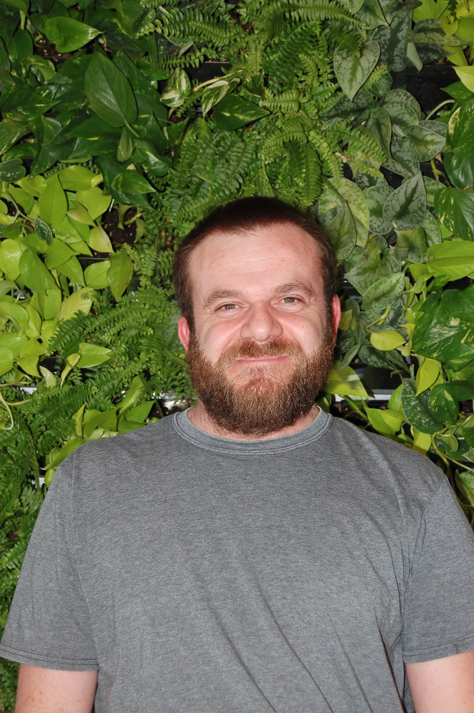 Un homme barbu souriant se tenant devant un mur de plantes vertes, comprenant des fougères et des feuilles de pothos.