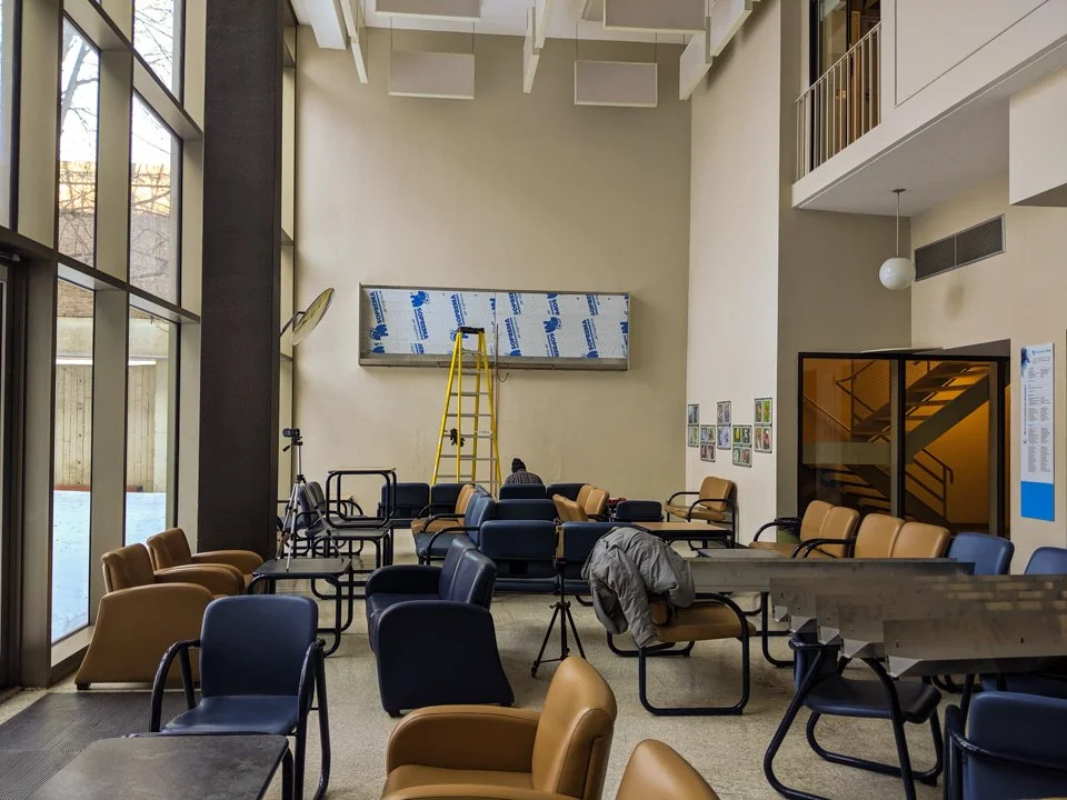 Une salle d'attente ou un espace de repos avec des fauteuils et des tables, une échelle jaune, un tableau blanc, et une personne assise en arrière-plan à côté d'un mur.