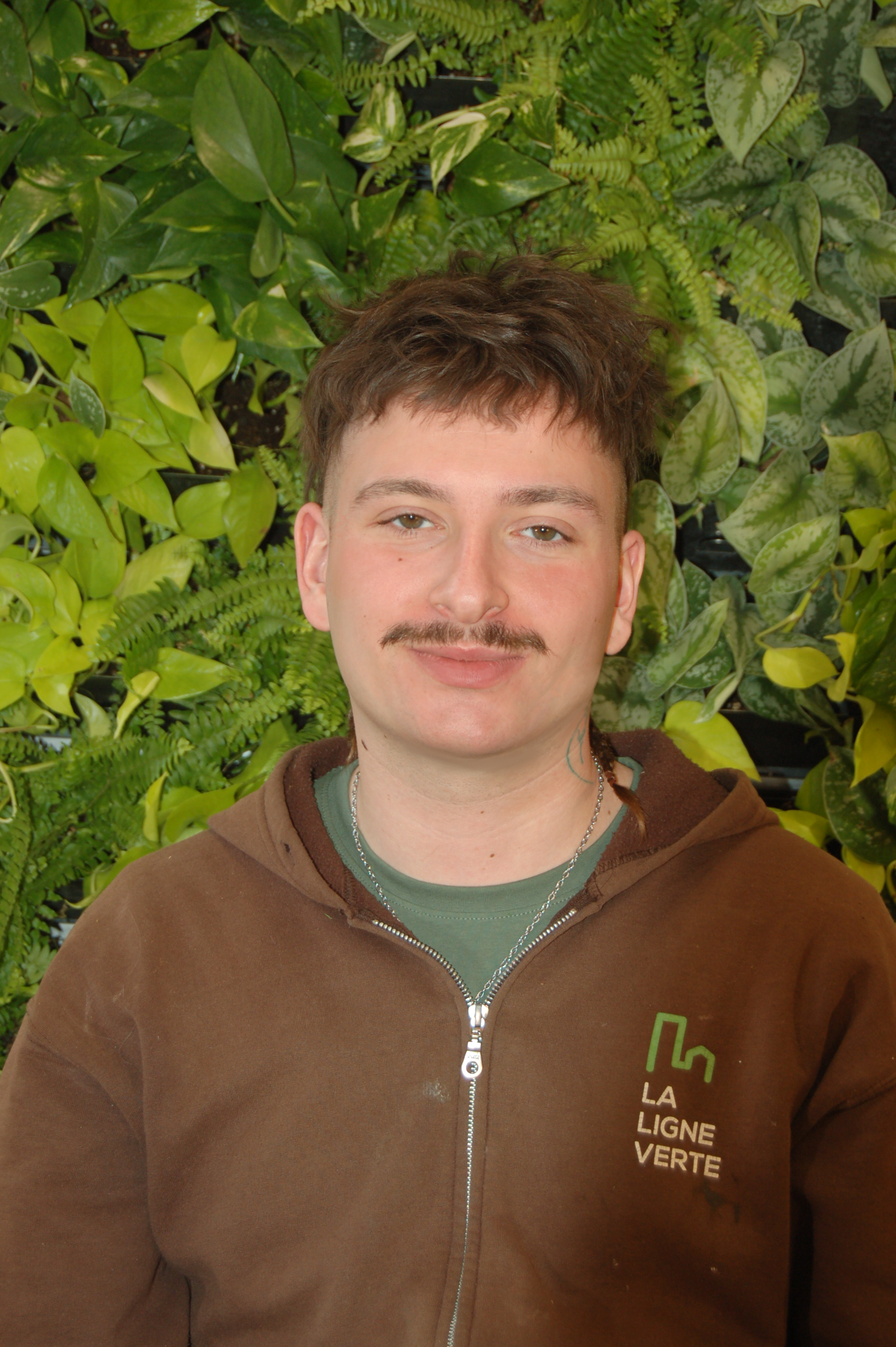Un jeune homme avec une moustache, portant une veste brune avec un logo vert et blanc, se tient devant un mur de plantes vertes.