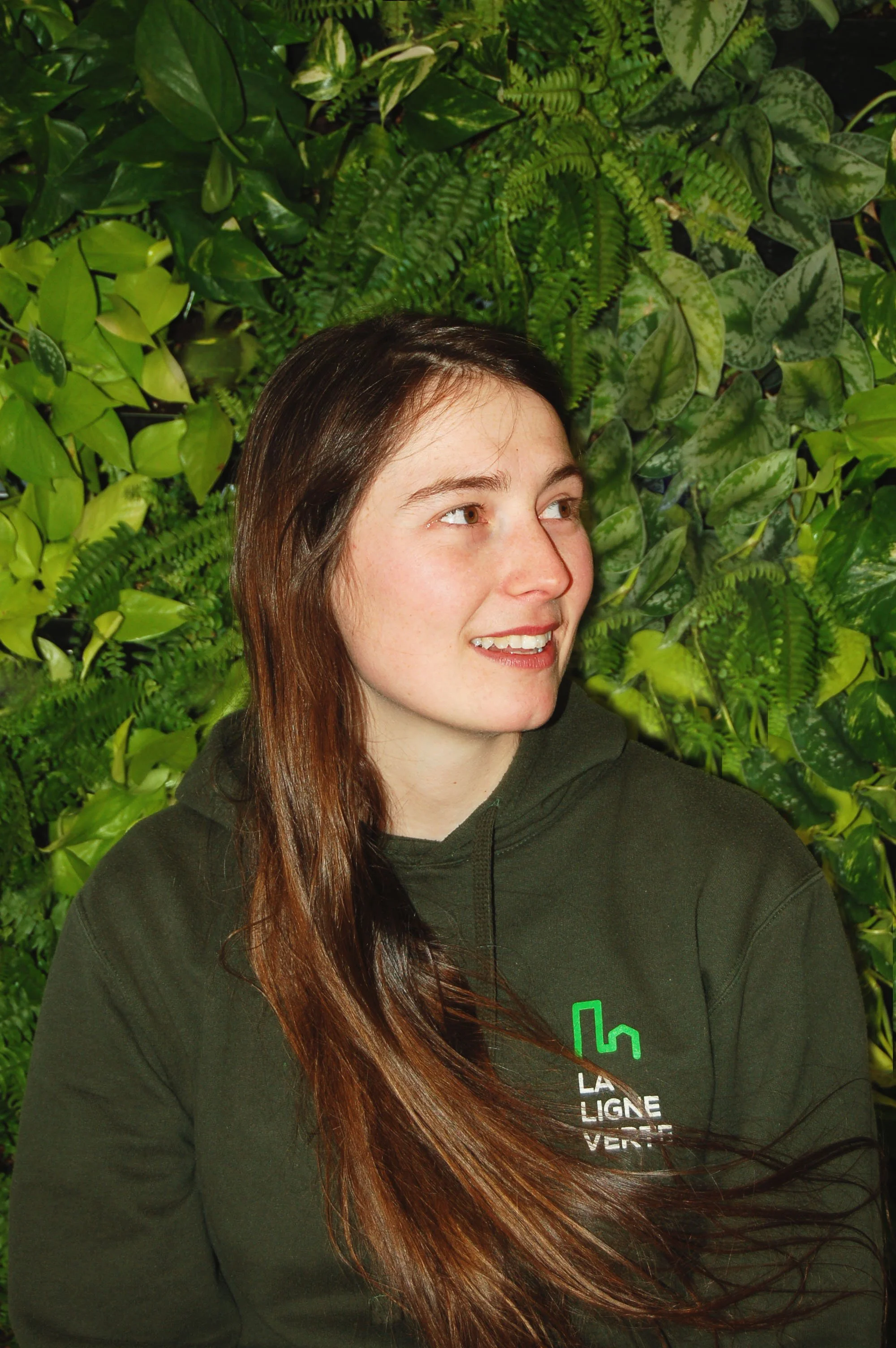 Jeune femme avec long cheveux bruns, portant un sweat à capuche vert, se tenant devant un mur de plantes verdoyantes.