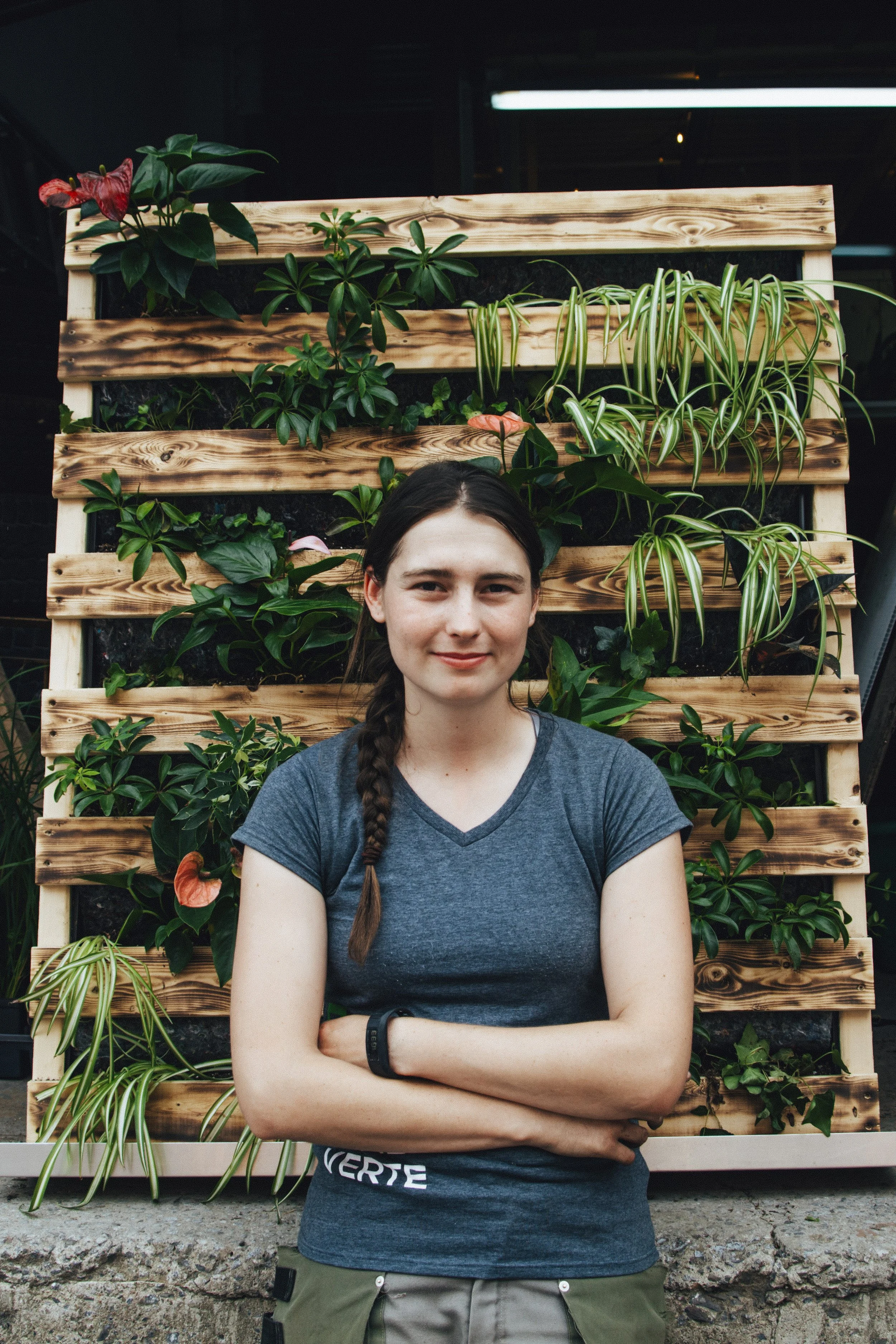 Jeune femme avec une tresse, debout devant un mur végétal en bois avec différentes plantes vertes.