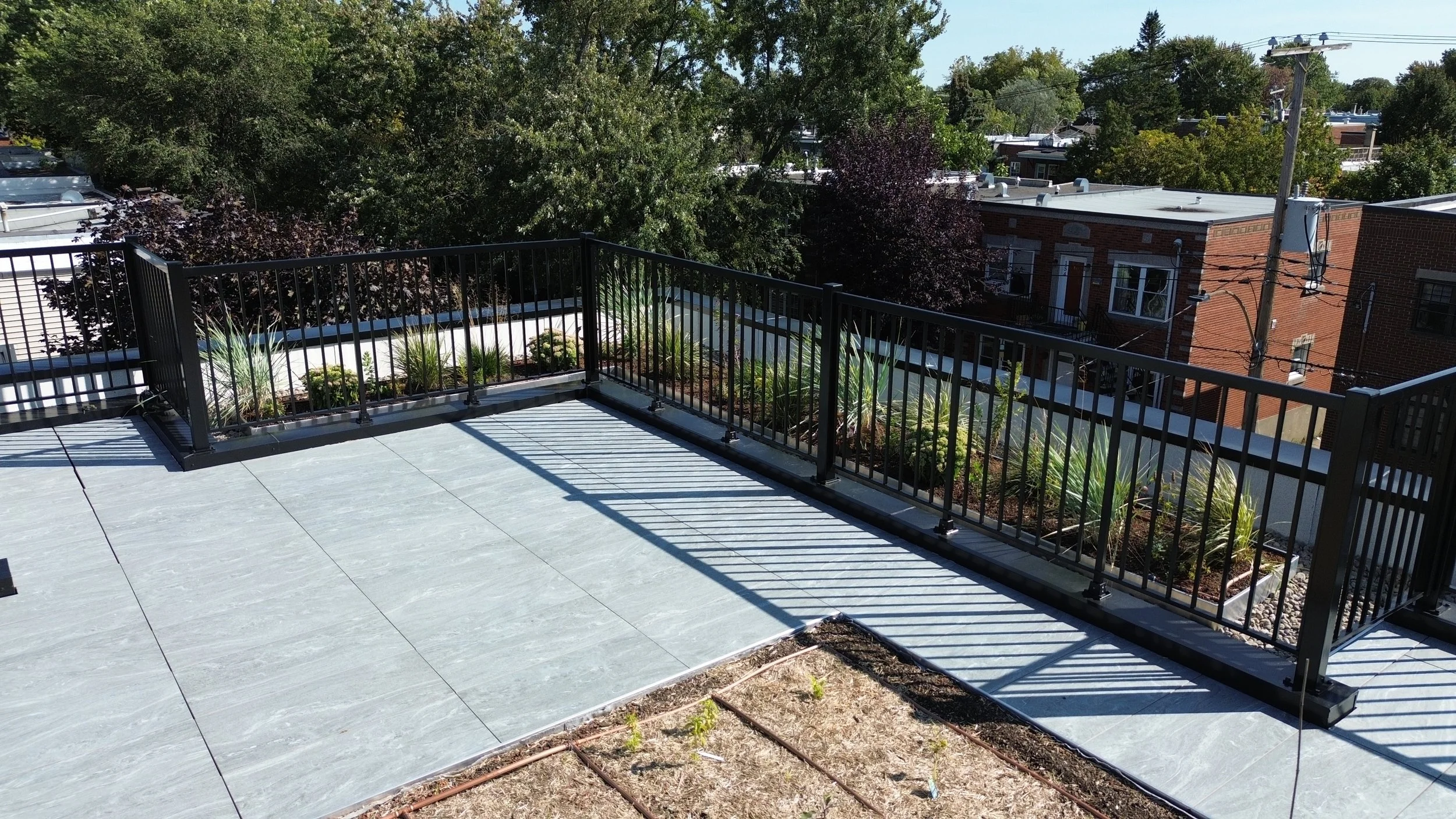 Terrasse en haut avec un sol en carrelage gris, une clôture en métal noir, et des plantes dans des jardinières. Vue sur des bâtiments résidentiels et des arbres en arrière-plan.