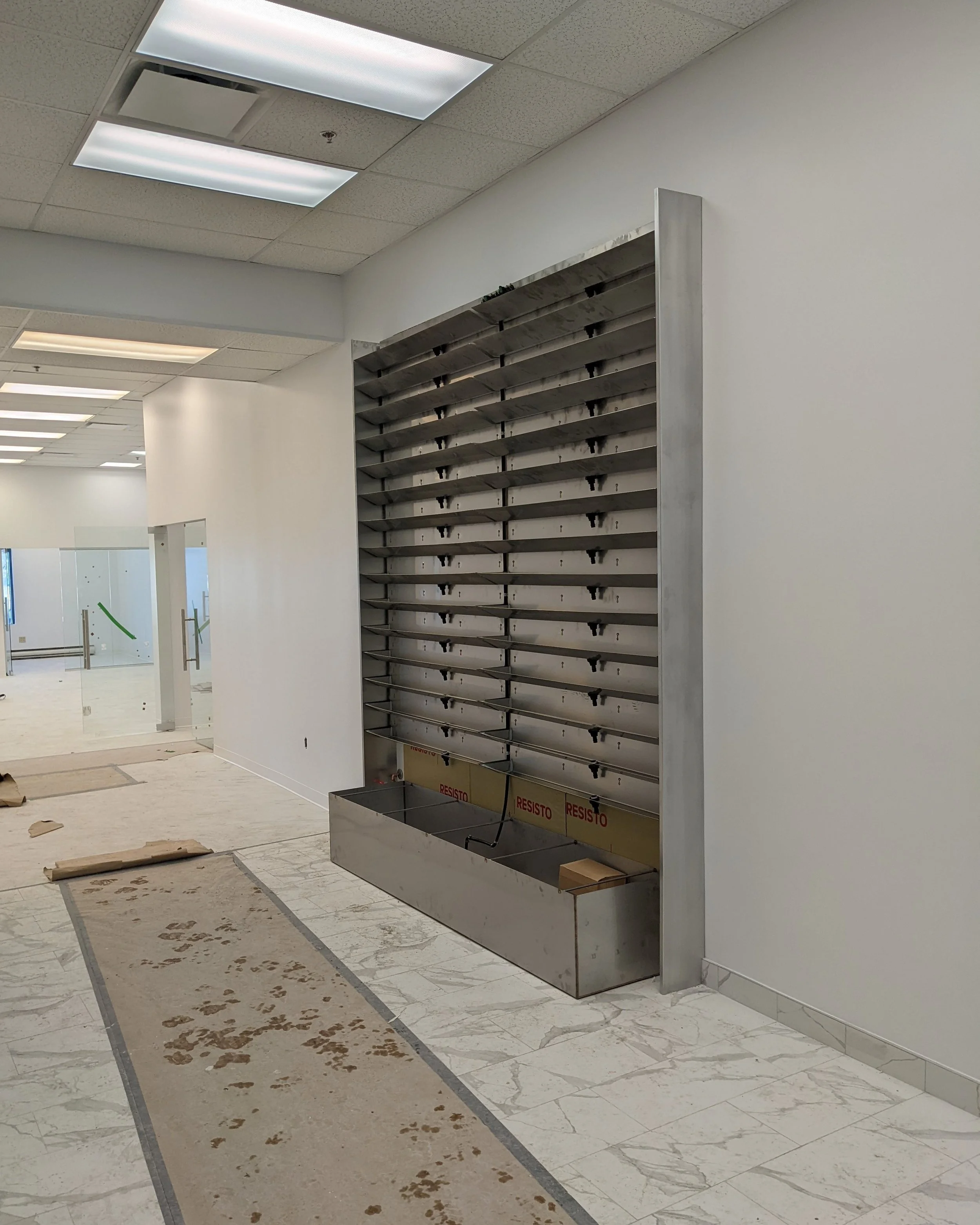 Intérieur d’un bâtiment en cours de construction ou de rénovation, avec un mur blanc, un plafond en dalles de plafond, et un mur métallique avec des étagères en acier, probablement pour des distributeurs ou des casiers.