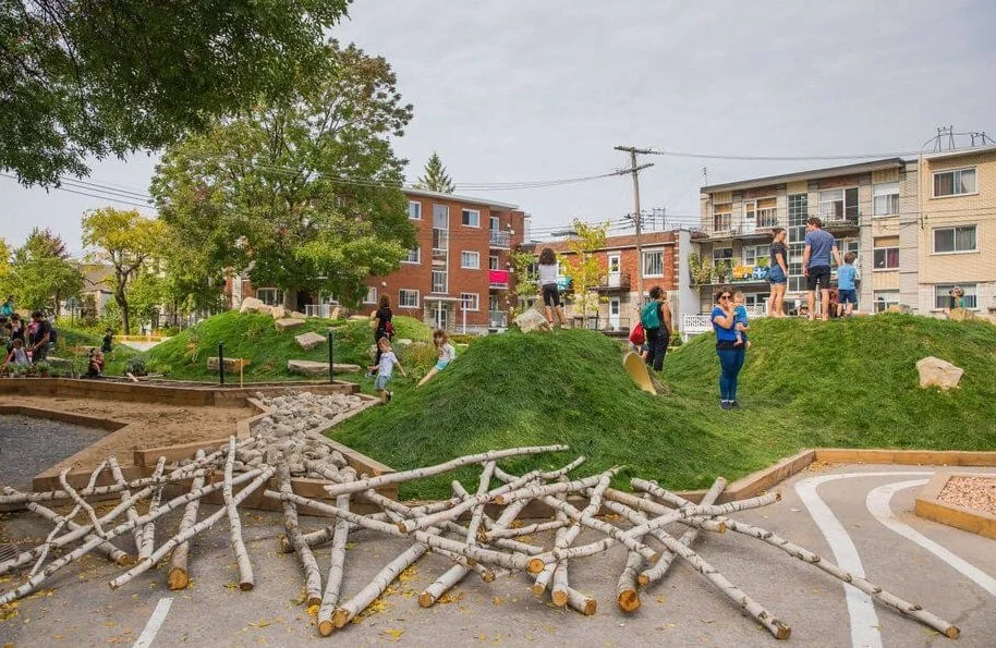 Un parc de jeux urbain avec des adultes et des enfants, avec des buttes herbeuses et des éléments en bois pour jouer, entouré d'immeubles résidentiels, en journée.