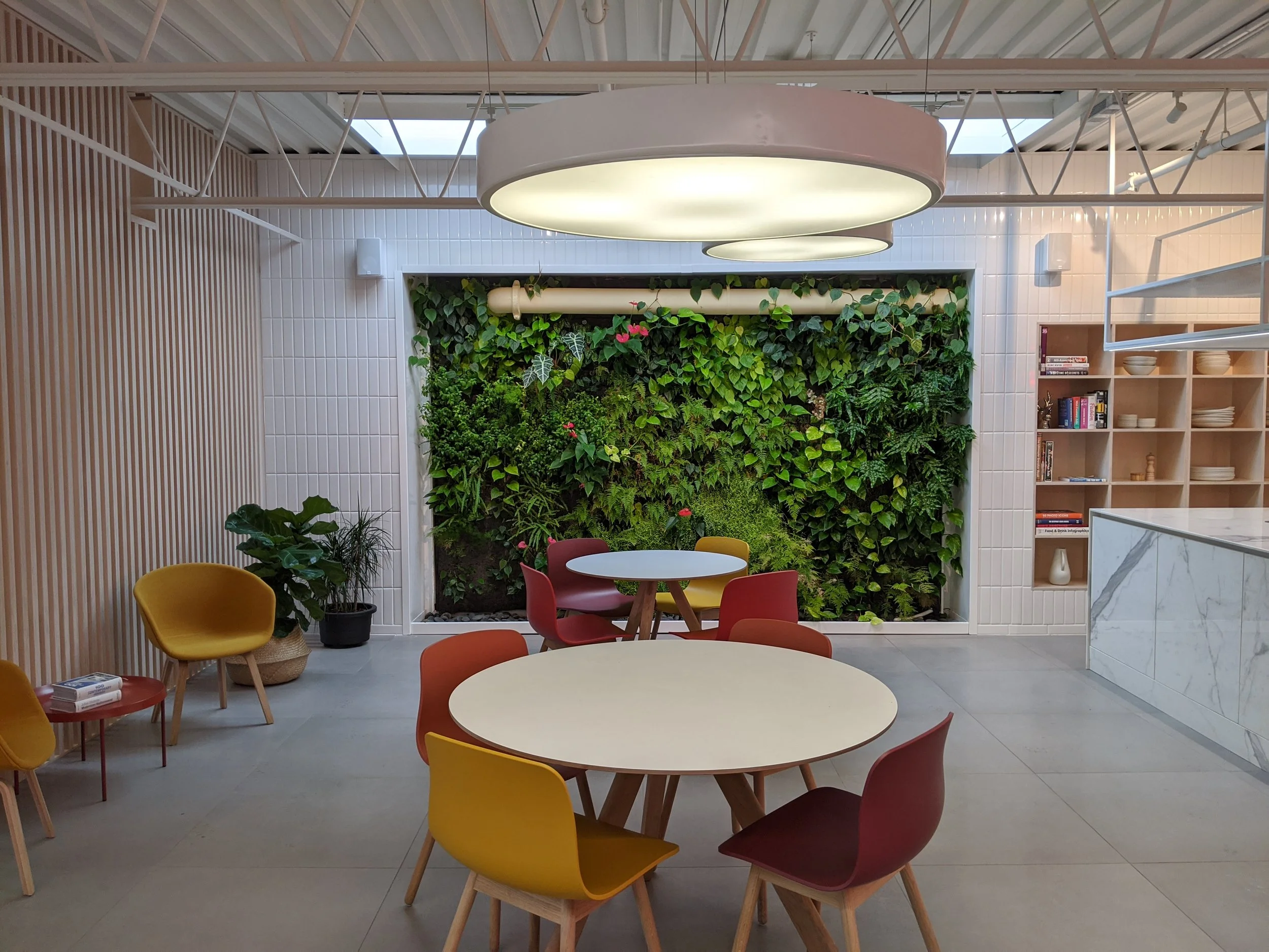 Intérieur d'un espace de restauration ou de coworking avec des tables rondes, des chaises colorées, un mur végétal en arrière-plan, des étagères avec des livres, et un comptoir en marbre blanc.