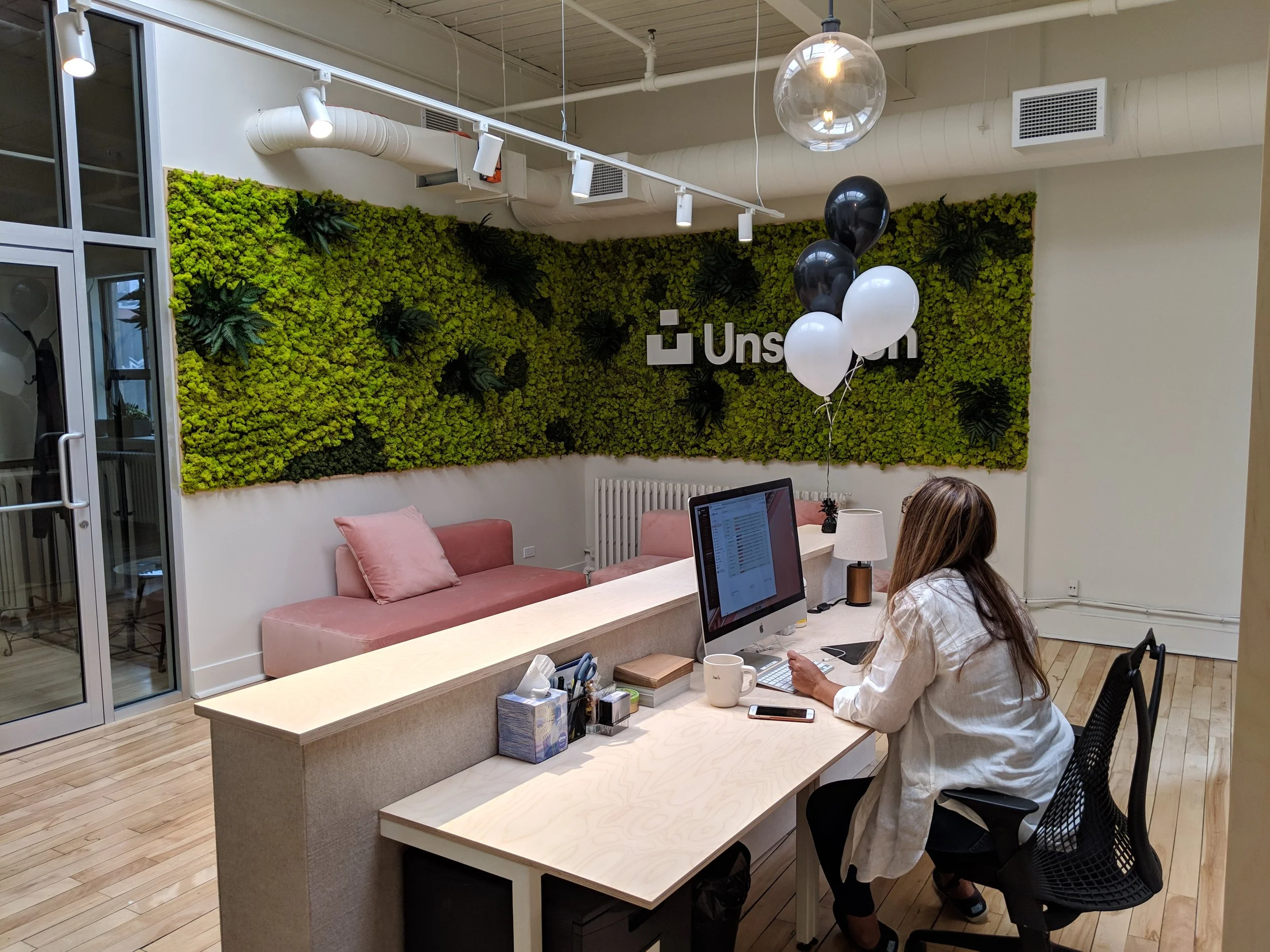Une femme travaille à son ordinateur dans un bureau moderne avec un mur végétal et un logo qui dit "Uns" en arrière-plan. Il y a des ballons noirs et blancs suspendus au plafond.