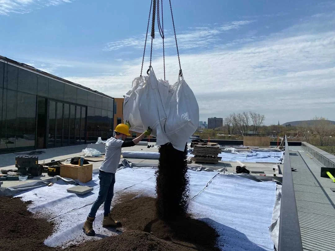 Un ouvrier portant un casque de chantier déverse un sac de gravier ou de terre sur un toit en construction.