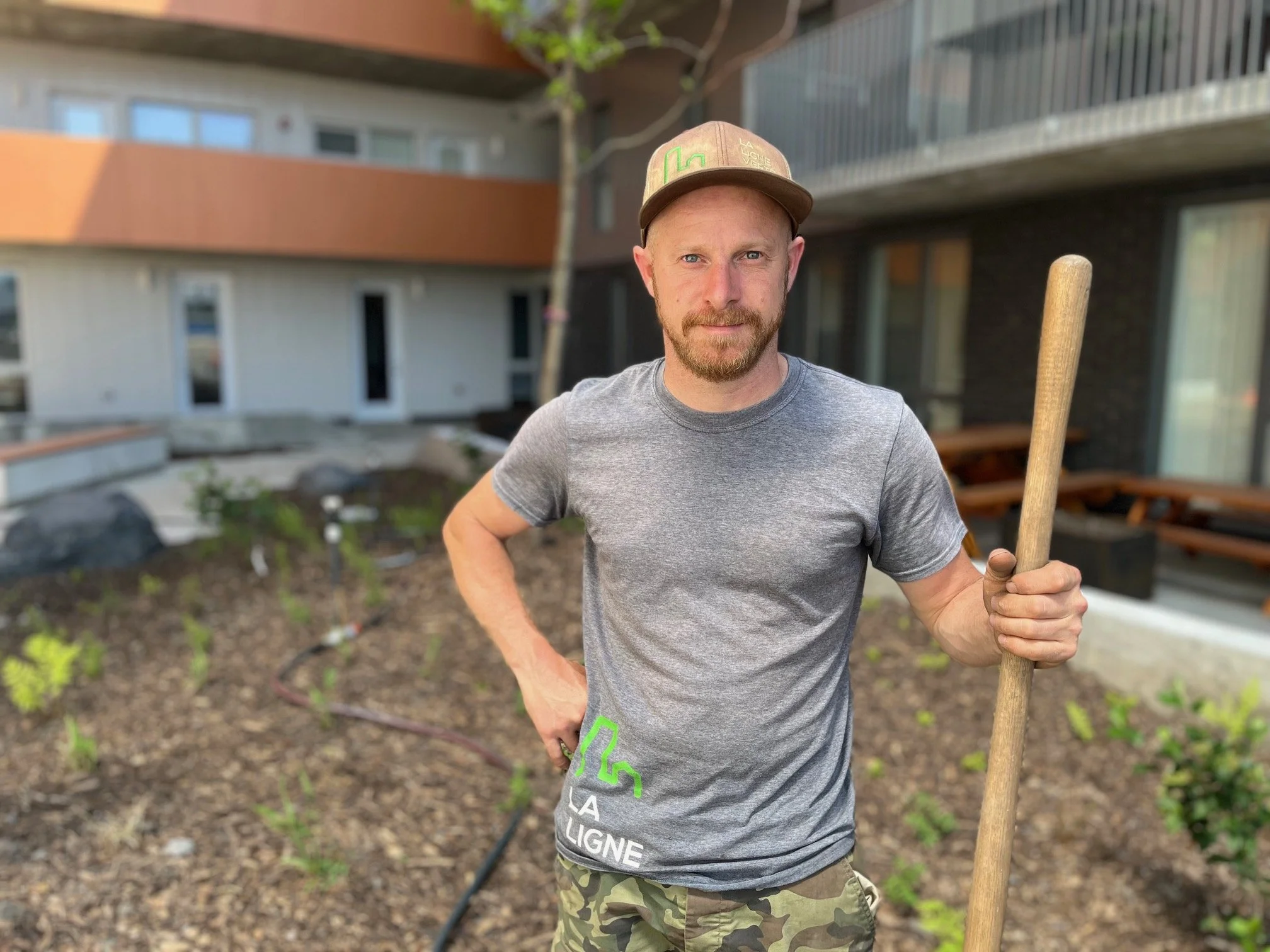 Un homme tenant une bêche, portant un t-shirt gris et un chapeau, dans un jardin.
