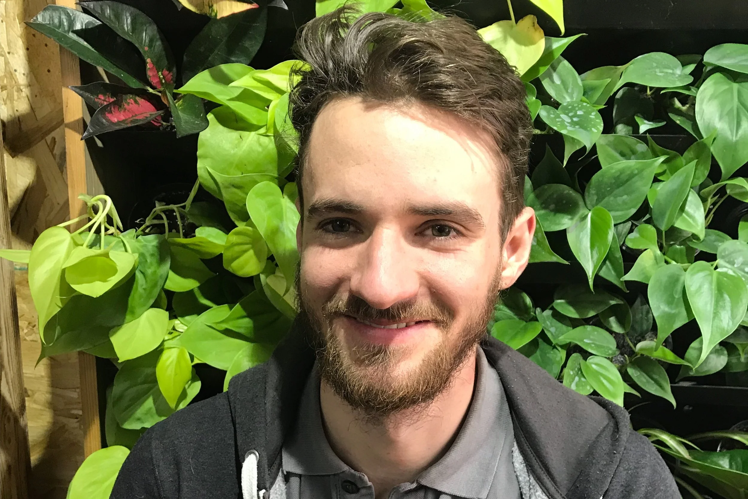 Un jeune homme souriant avec une barbe, porte un sweat noir et un polo gris, se trouve devant un mur de plantes vertes luxuriantes.