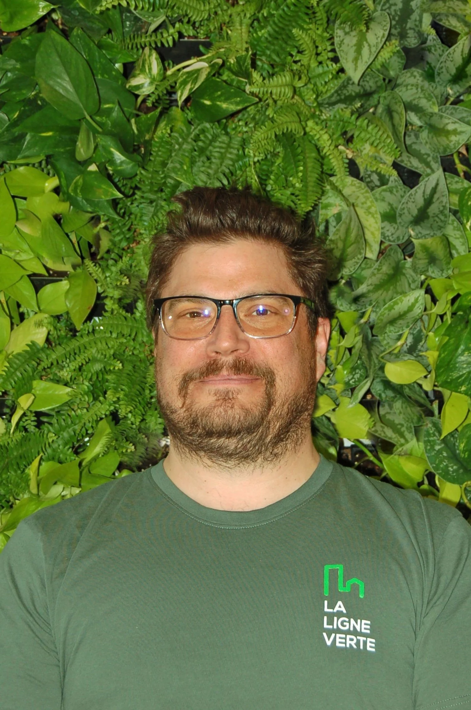 Un homme à lunettes et barbe, portant un t-shirt vert avec le logo 'La Ligne Verte', pose devant un mur de plantes vertes.