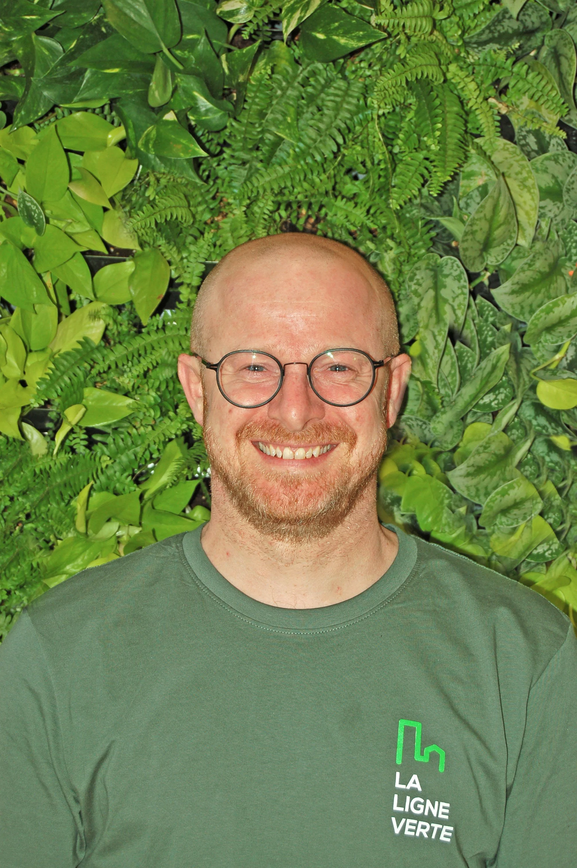 Un homme souriant portant un t-shirt vert avec le logo de La Ligne Verte, devant un mur de plantes vertes.