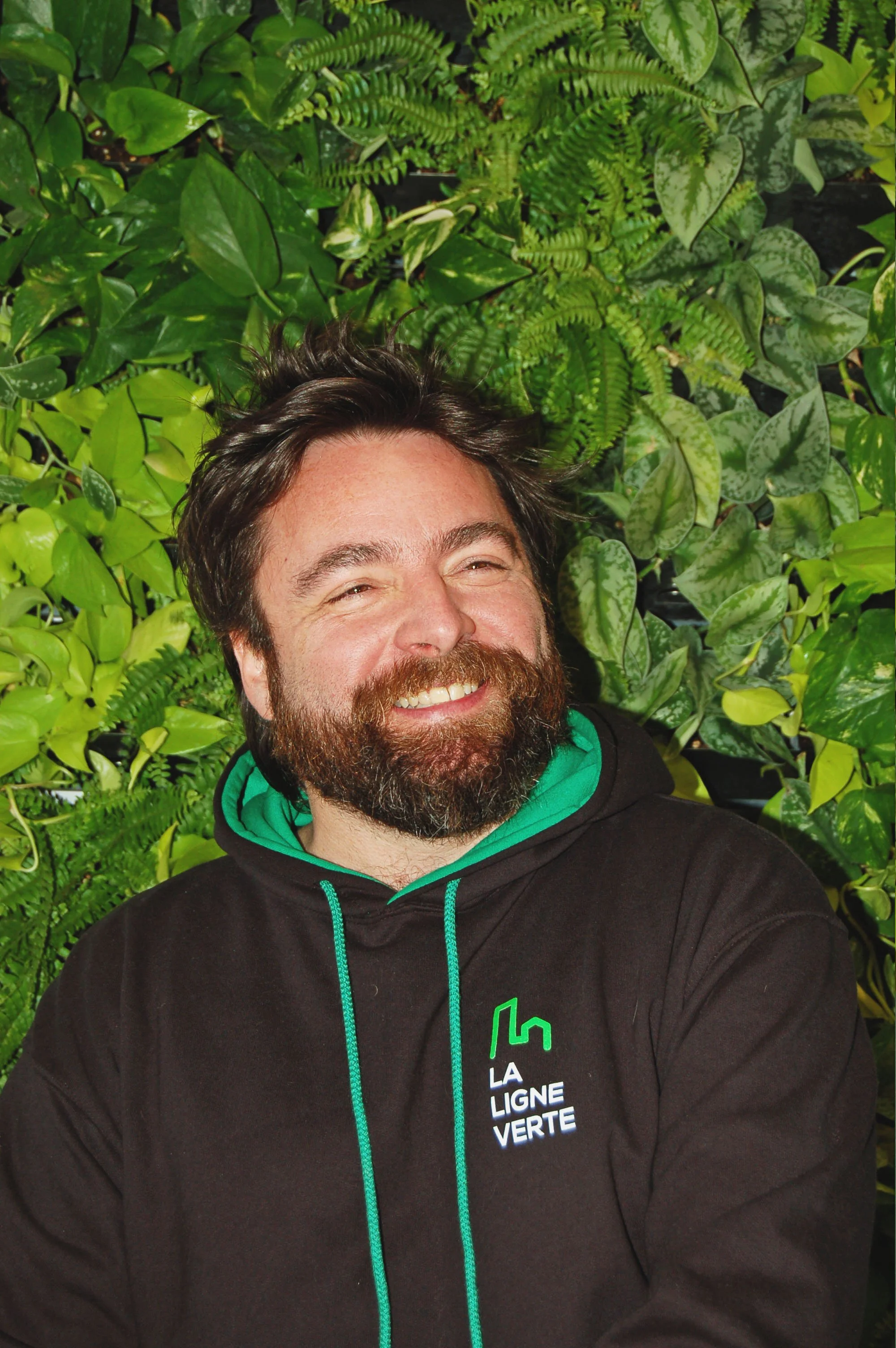 Un homme souriant avec une barbe, portant un sweat à capuche noir avec un logo vert et blanc et un fond de plantes vertes luxuriantes.