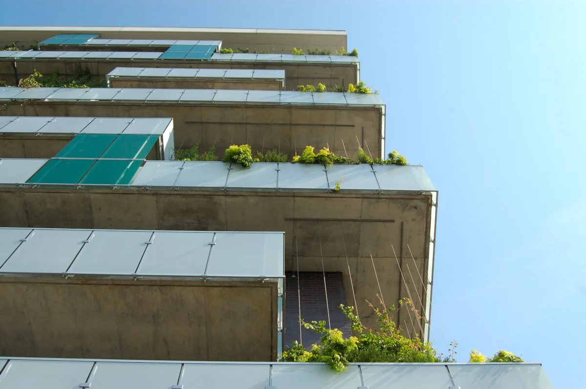 Vue en contre-plongée d'un bâtiment moderne avec des balcons en verre et des plantes vertes sur chaque étage, sous un ciel clair.