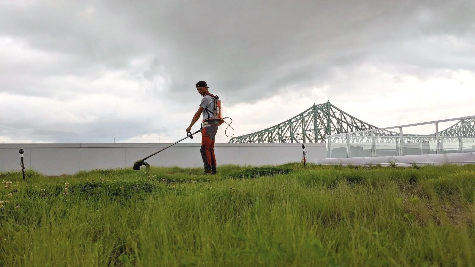 Un homme utilise une tondeuse à gazon sur une pelouse verte, avec un ciel nuageux et un pont en fer en arrière-plan.