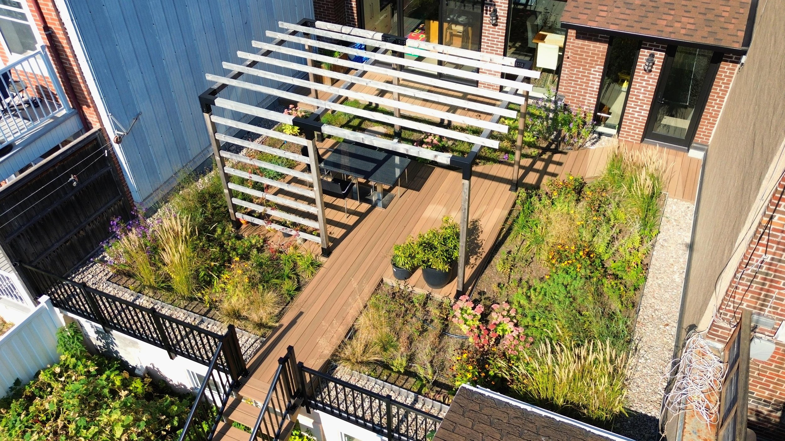 Une cour de jardin avec une terrasse en bois, une pergola en bois avec des plantes, un mur en briques, des plantations fleuries, et une table de pique-nique noire.