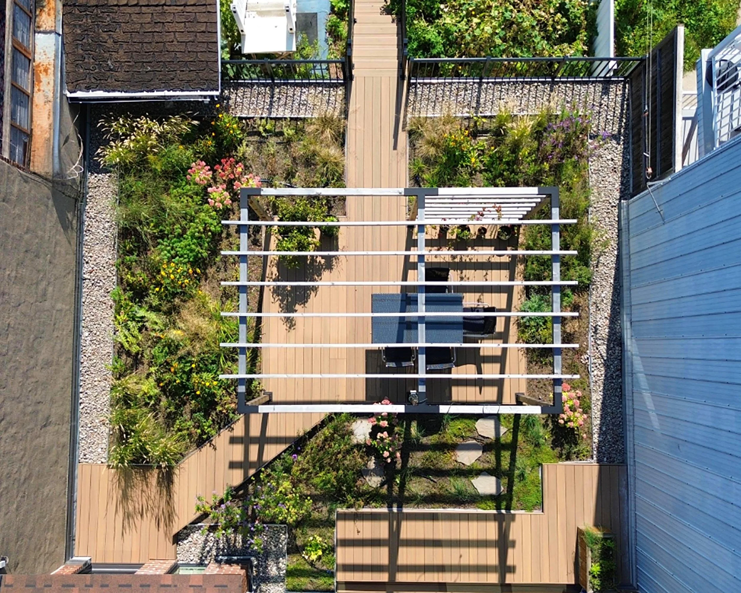 Vue aérienne d'un petit jardin en terrasse avec une pergola en bois, des plantes et des fleurs, un coin salon avec une table et des chaises, entouré de clôtures et bâtiments.