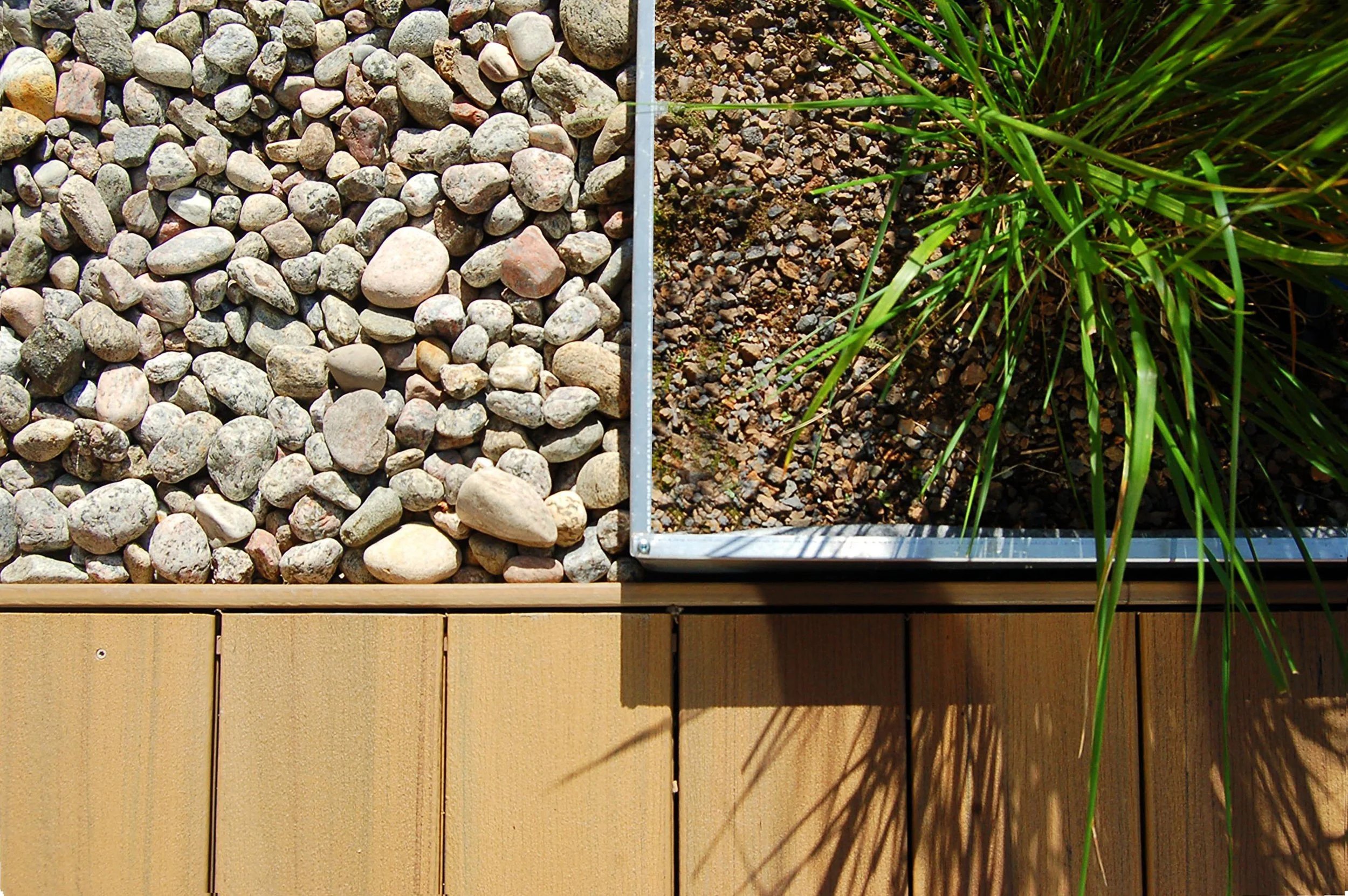 Division en deux sections de différentes textures : la gauche avec des pierres de différentes tailles, la droite avec de la terre et une plante verte, séparées par une bordure métallique, en dessous d'une structure en bois.