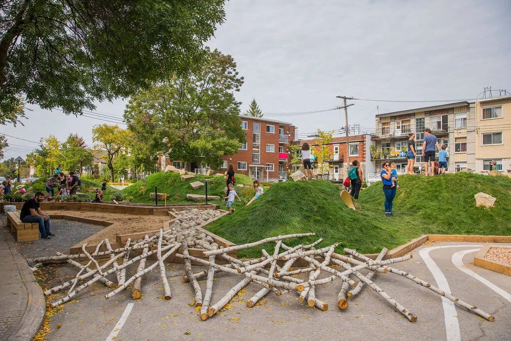 Parc de jeux avec des enfants et adultes, arbres, buttes de terre et bois, et un bâtiment résidentiel à l'arrière