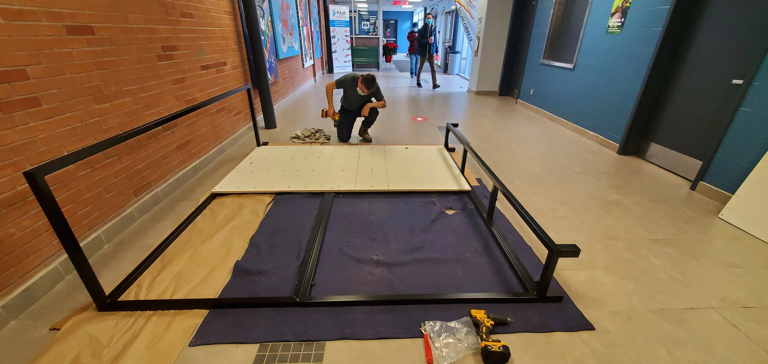 Installation d'une plateforme ou d'un meuble en bois dans un centre commercial ou un établissement public, avec deux personnes en train de travailler, une avec un tournevis et des outils, dans un environnement intérieur moderne.
