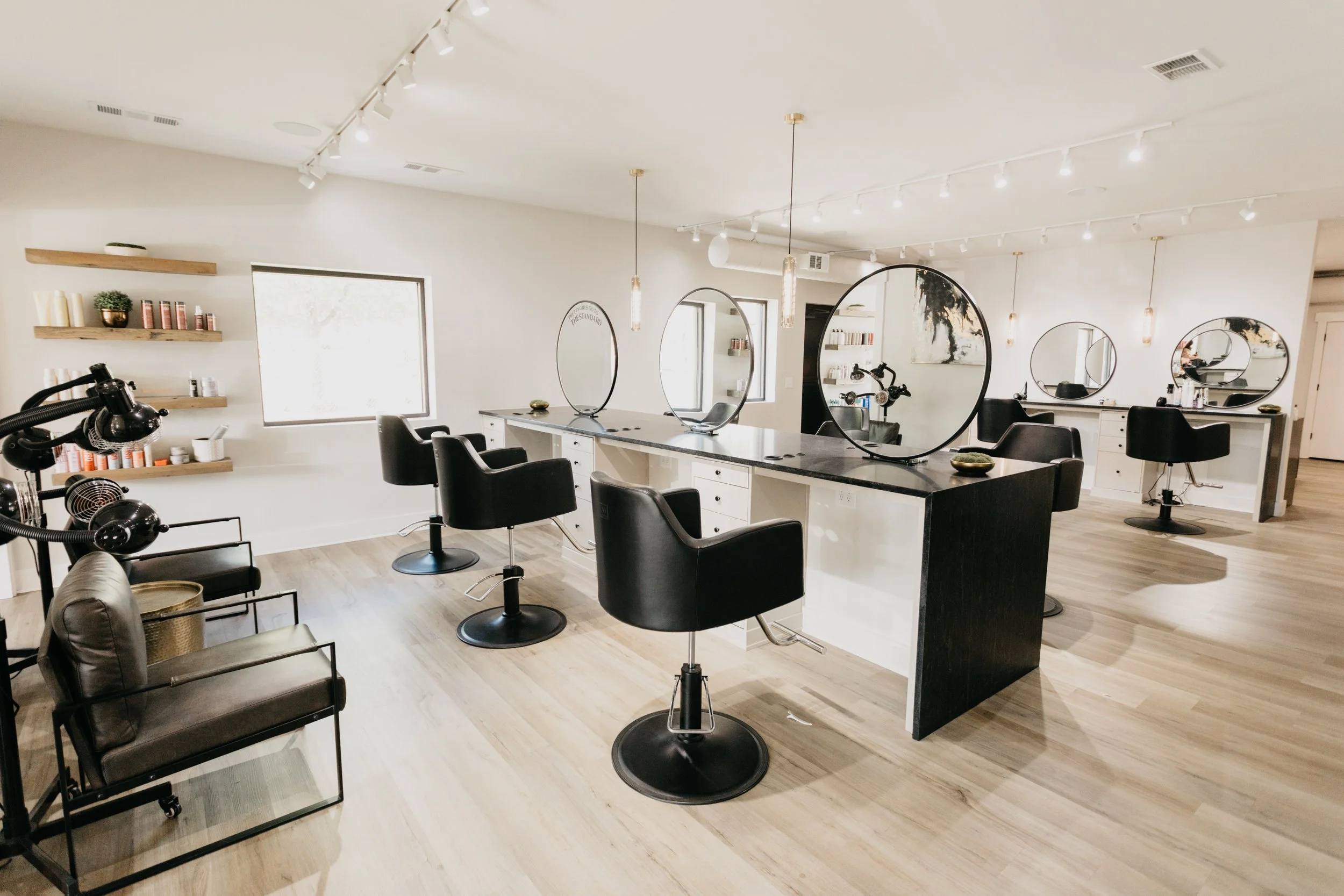 A modern hair salon with black styling chairs, large mirrors with circular frames, and overhead lighting. Shelves with hair products and accessories are on the wall, and a customer awaiting service is seated in a vintage-style brown chair. The salon has light-colored wooden flooring and a clean, minimalist design.