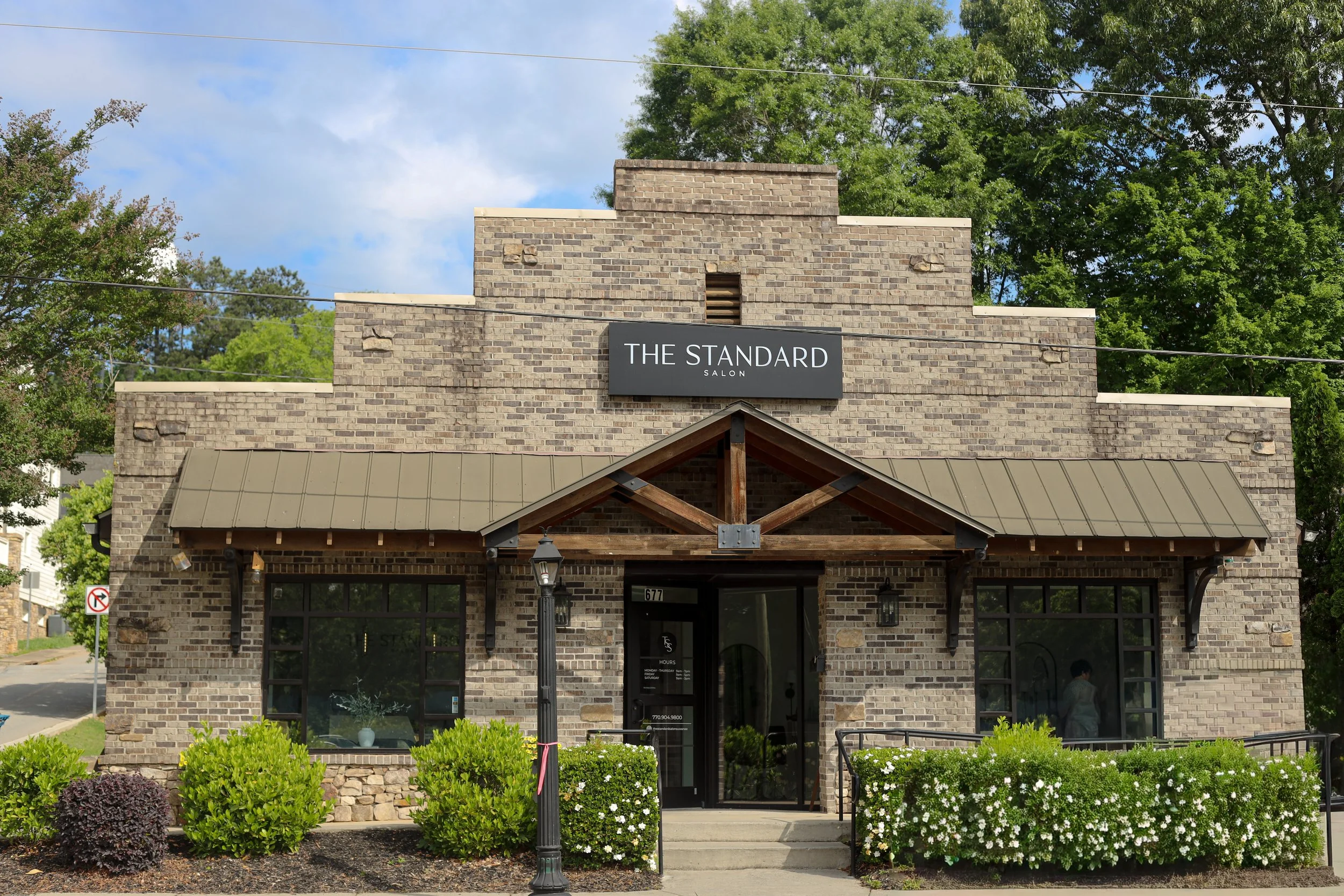 The storefront of 'The Standard' salon, a brick building with a sign on it, surrounded by greenery, flowers, and trees on a sunny day.