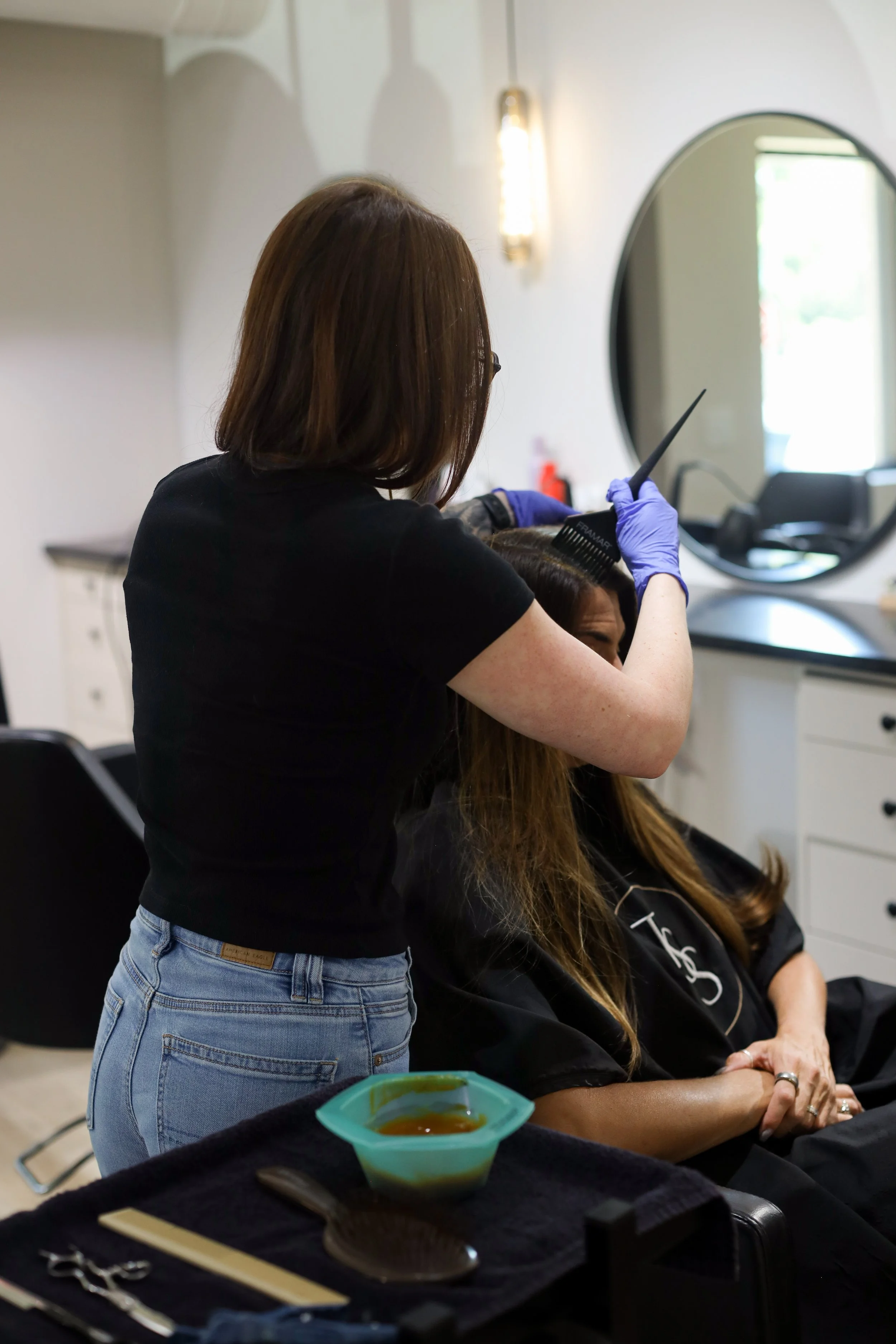 A woman with long brown hair wearing a white tank top and blue jeans applying hair dye to a young girl's blond hair who is sitting in a black salon chair, with foil in her hair.