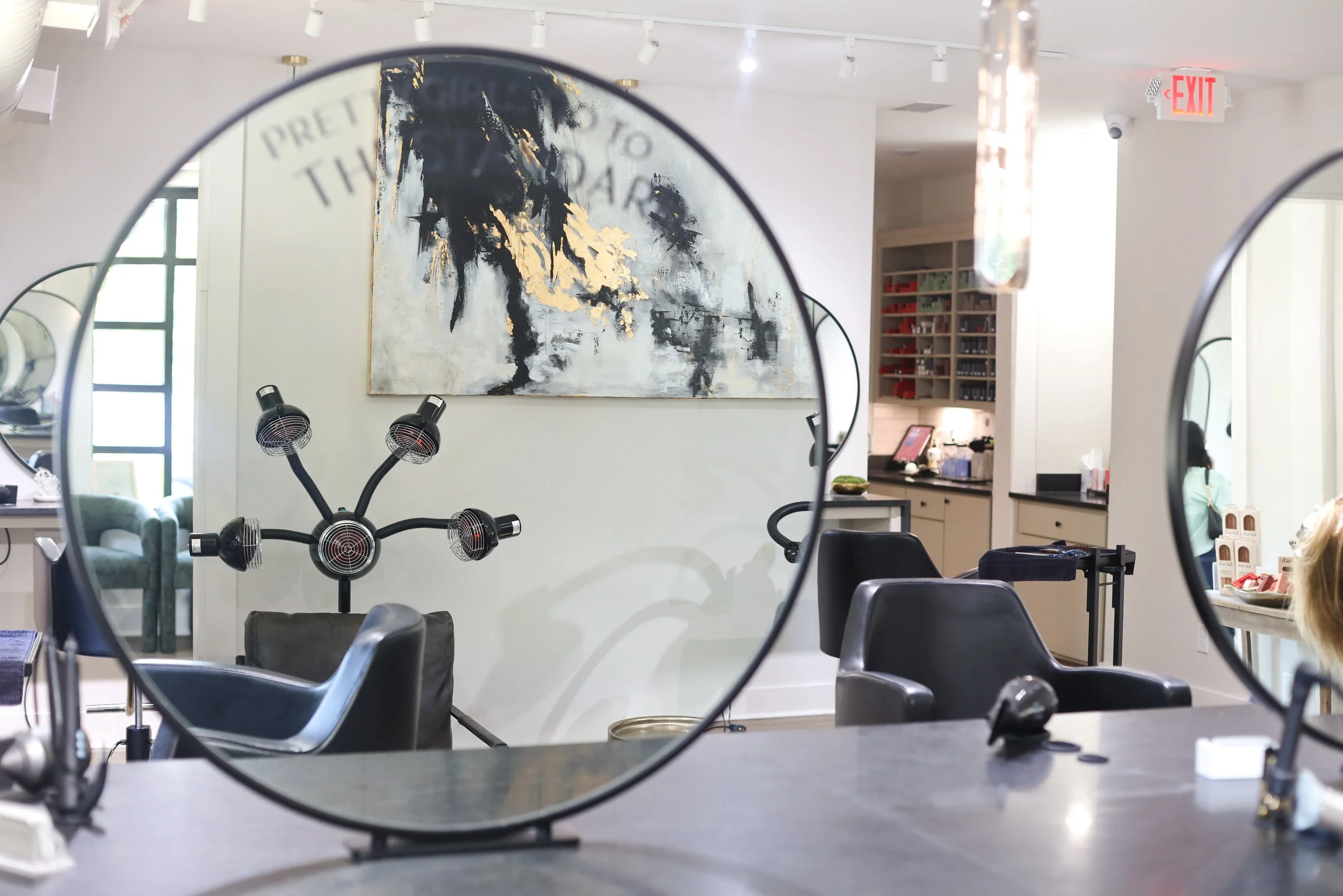 Interior of a salon or barber shop viewed through two round mirrors showing a large abstract portrait painting on the wall, salon chairs, hair styling tools, and a customer in the background.