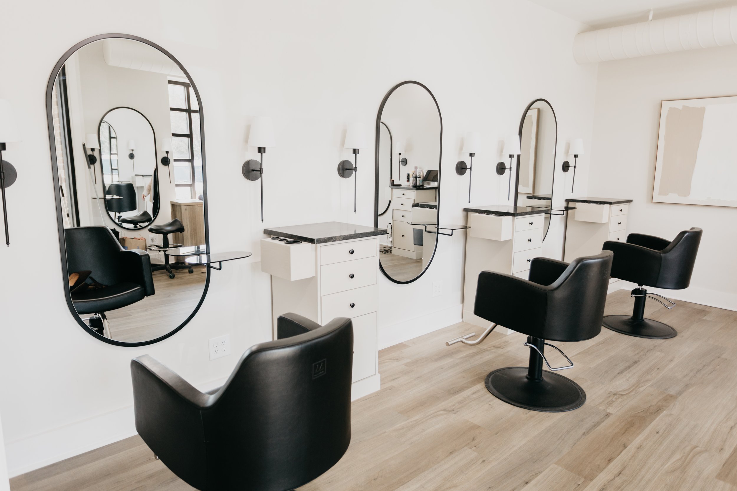 Modern salon with white walls, black wall-mounted lamps, tall mirrors, black chairs, and minimal decor.