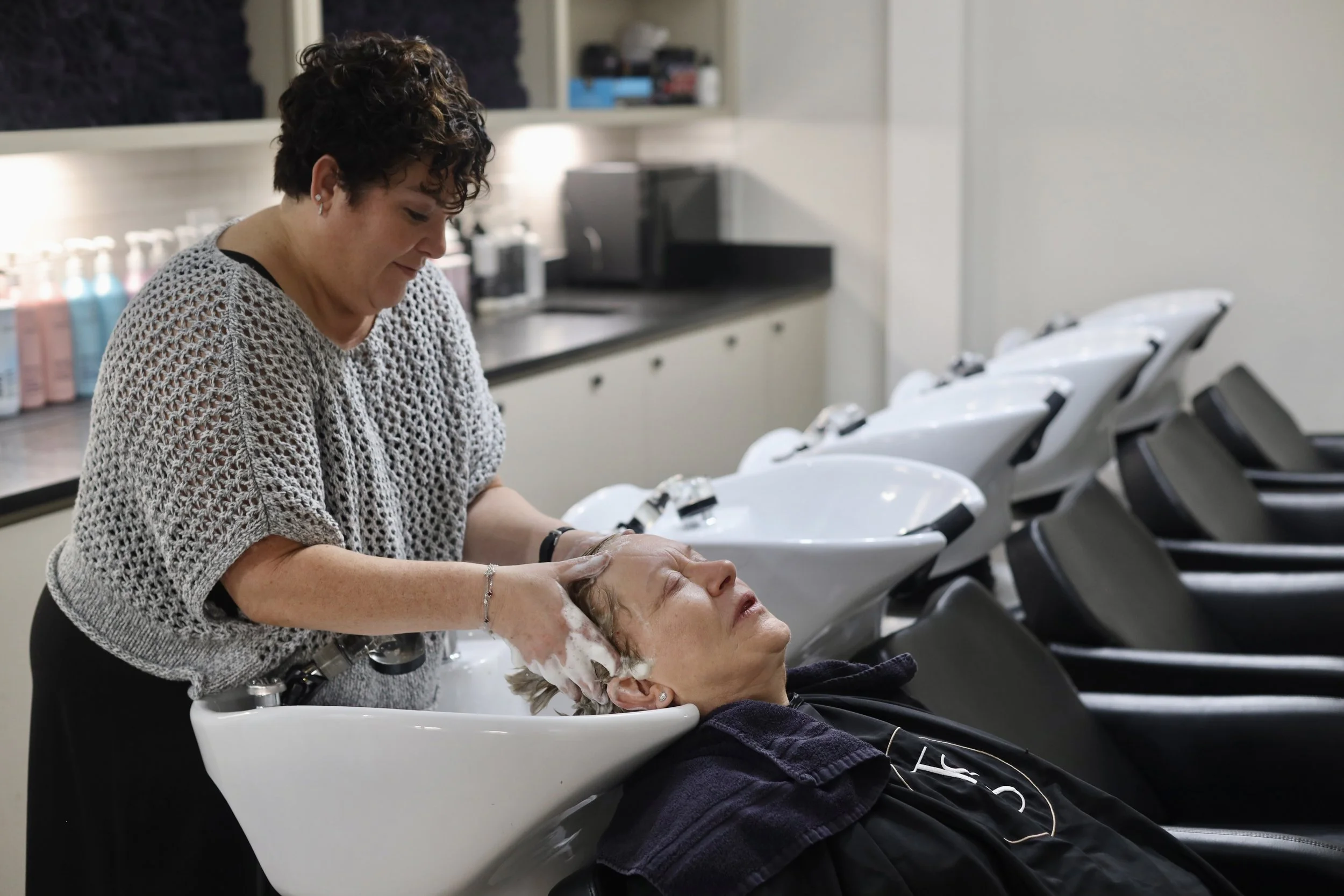 A woman receiving a hair wash at a salon, lying back with her head in a sink while another woman washes her hair.