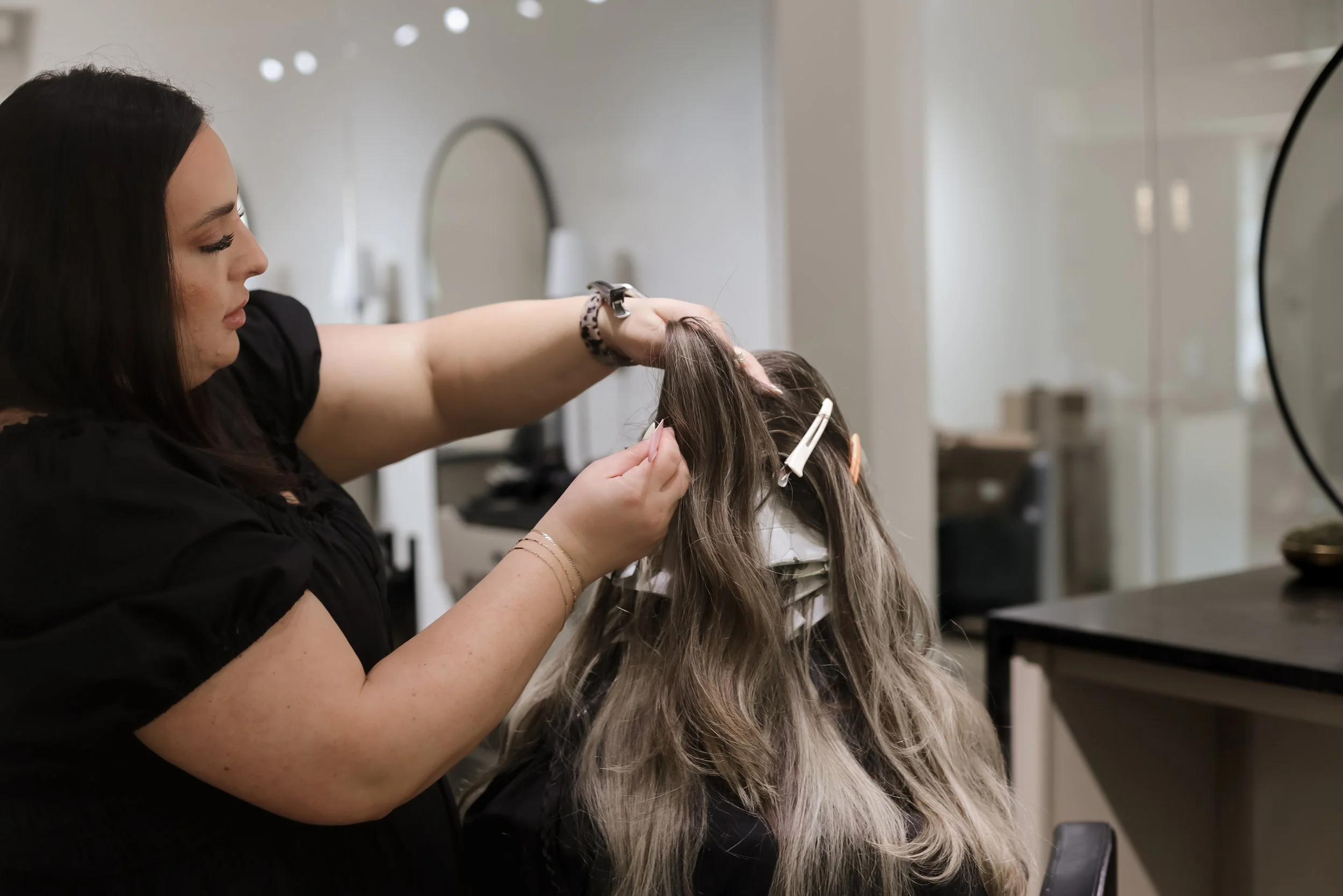 A woman with dark hair getting her hair styled or cut at a salon by a stylist. The stylist is touching the woman's hair.