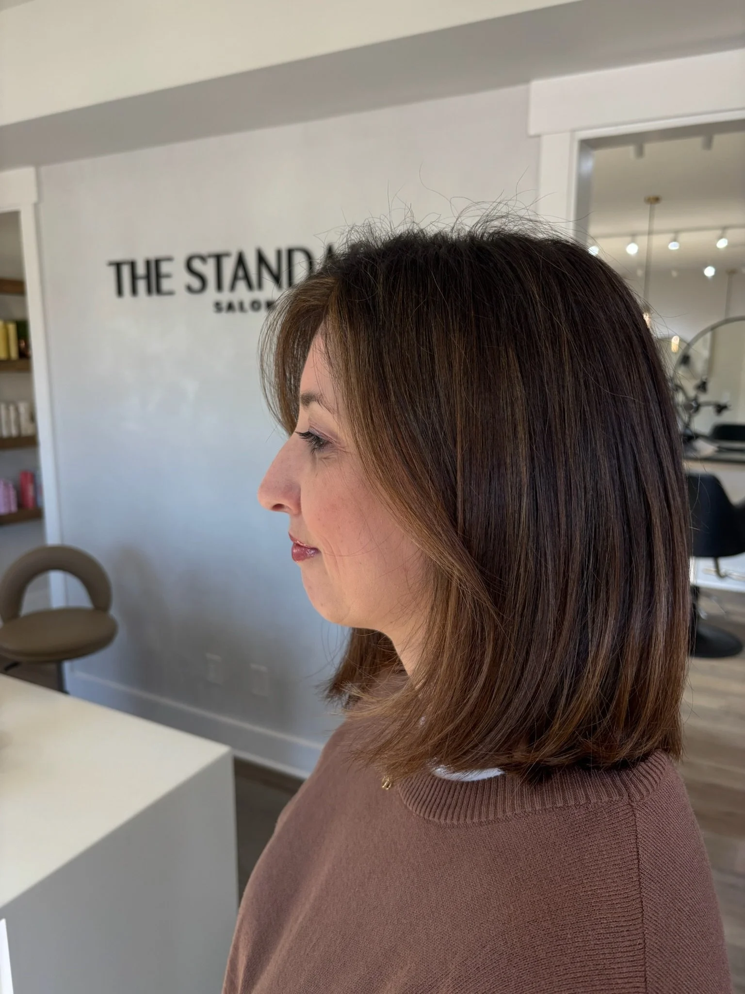 A woman with layered brown hair with blonde highlights, wearing a brown sweater, is inside a salon. The salon has a sign that reads 'The Stand' on the wall, with shelves of hair products and a salon chair visible in the background.