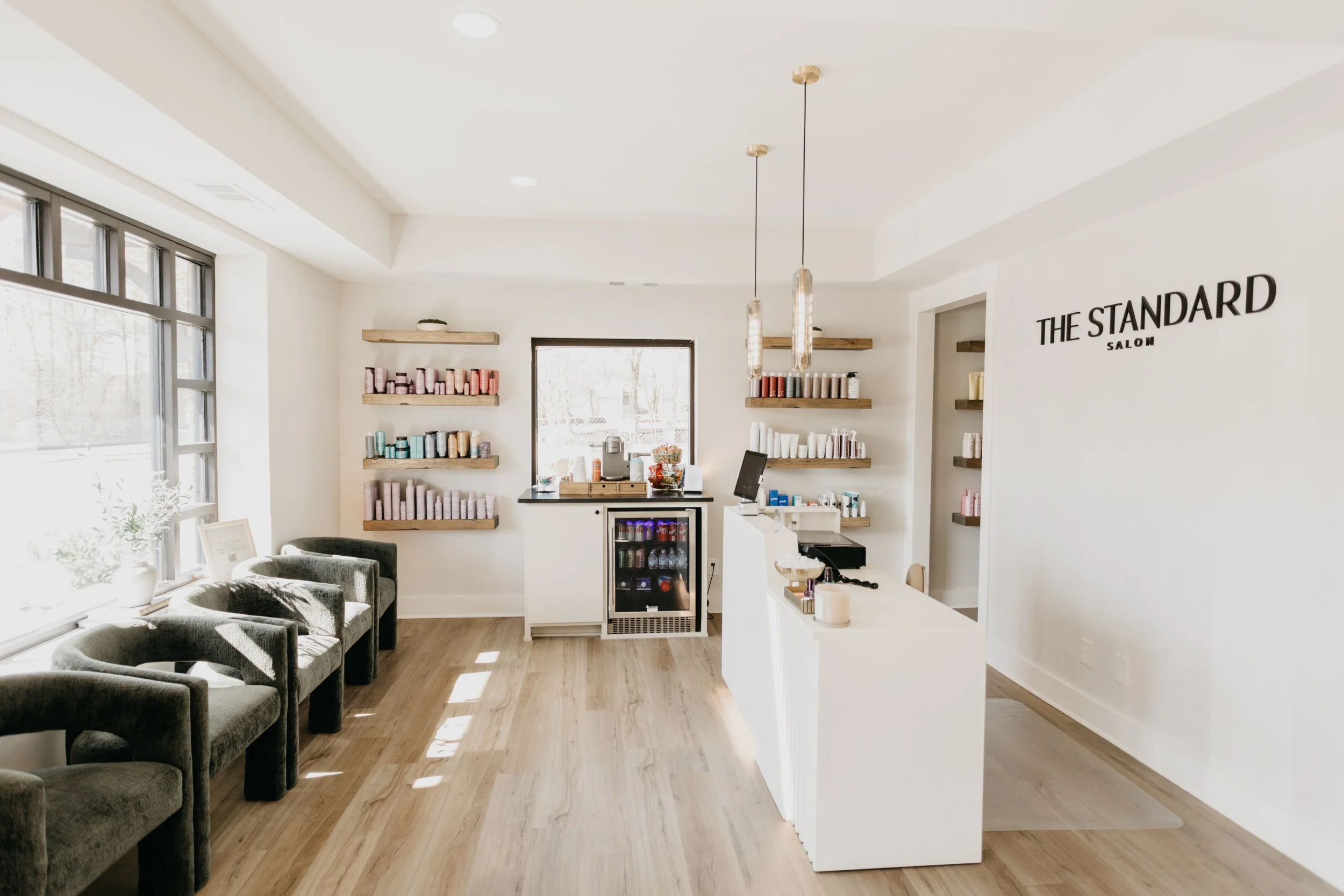Interior of a modern salon with white walls, wooden flooring, and natural light from large windows. The space features a reception counter, shelves with hair products, and seating for clients.