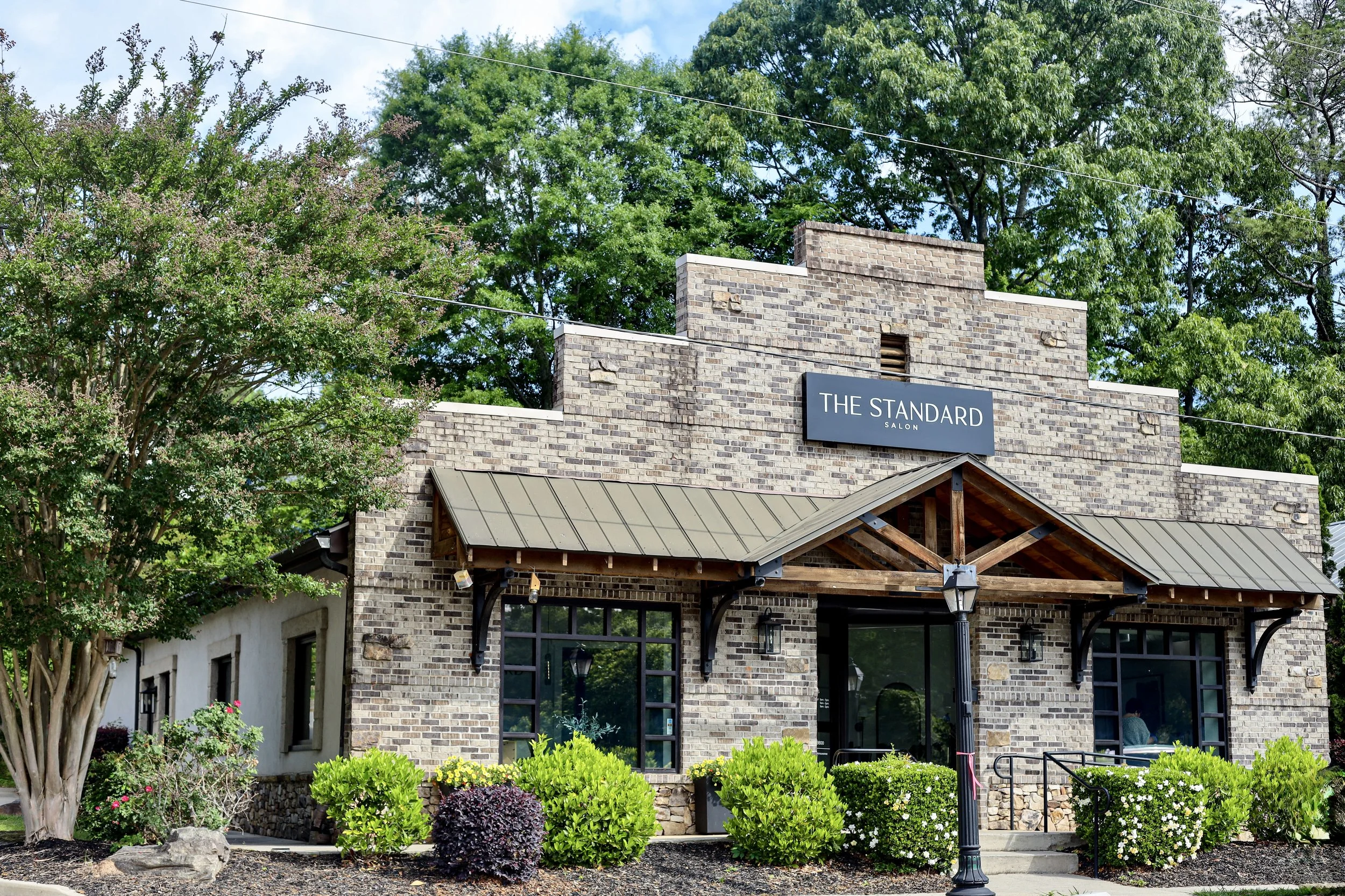 Brick building with a sign that says 'The Standard Salon', large windows, a metal awning, and surrounded by greenery including shrubs and trees.