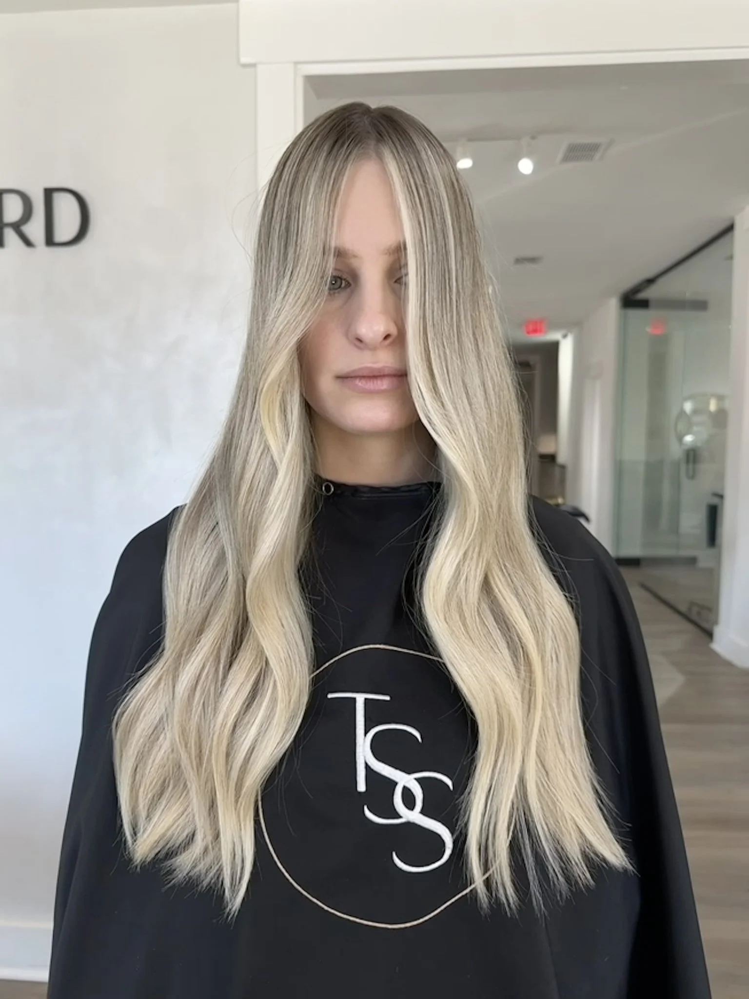 A woman with long, wavy blonde hair with darker roots, standing in a modern salon interior.