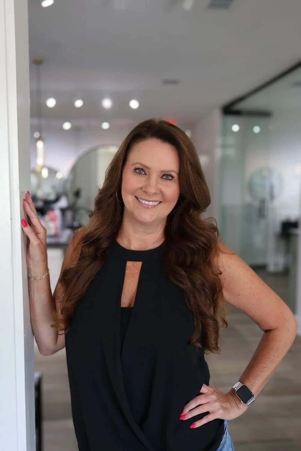 A young woman with long, wavy brown hair and a white top standing indoors near a wall, with a mirror and salon or studio background.