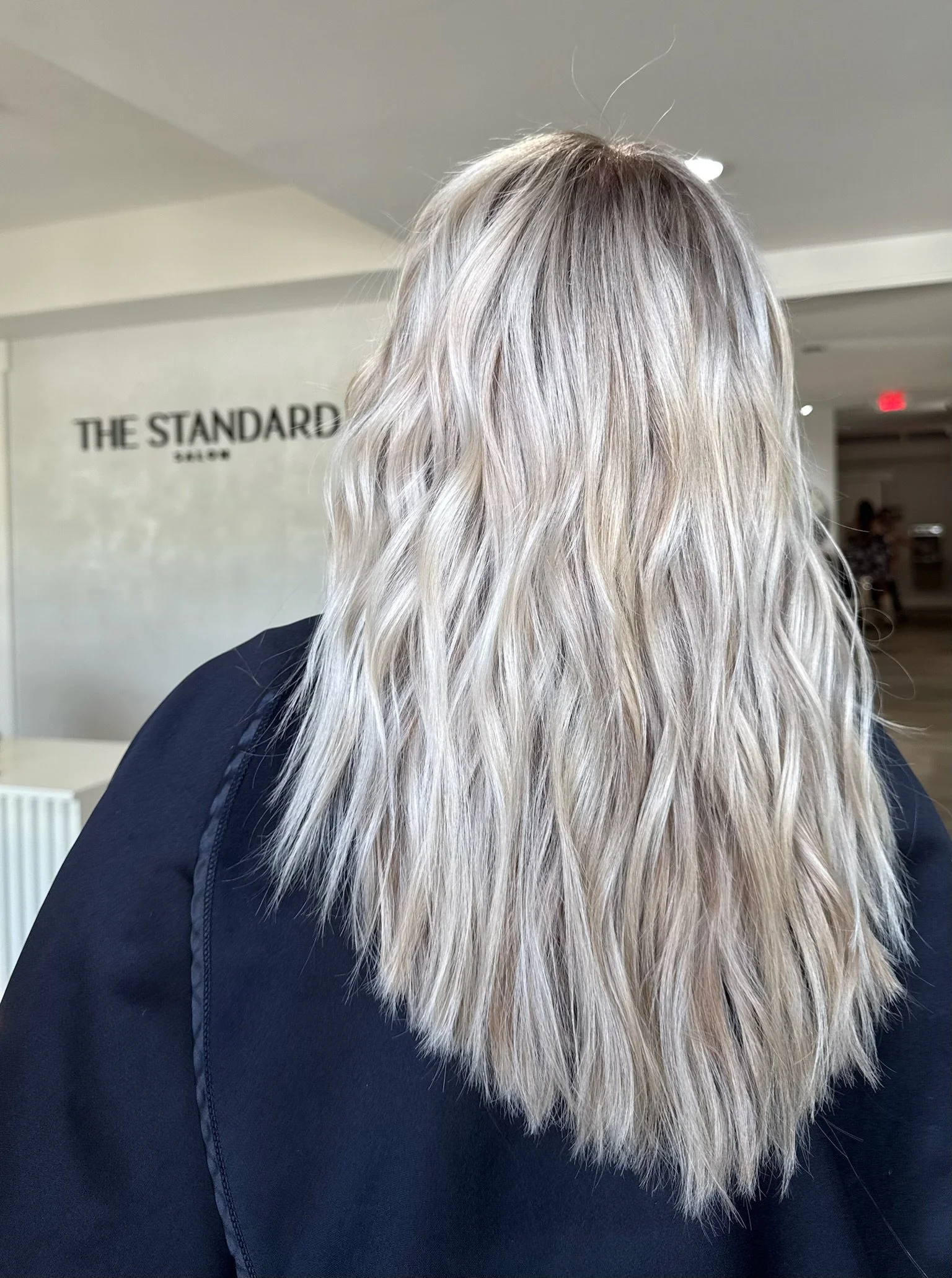 Back view of a person with long, wavy, platinum blonde hair wearing a black top, standing in an indoor setting with a sign that reads 'THE STANDARD NYC' in the background.