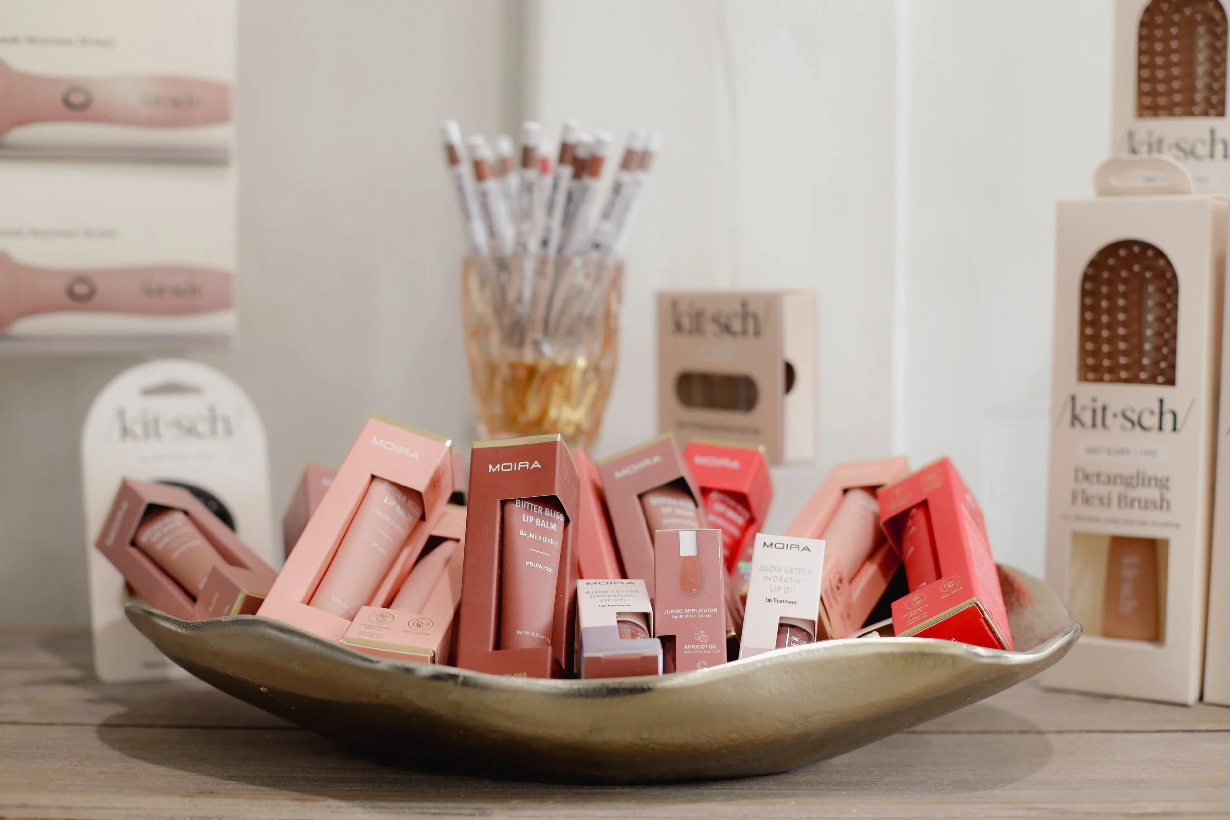 A decorative bowl filled with various lip balm products in pink and beige packaging. In the background, there are boxes and containers labeled 'kit-sh' and 'Moira' on a store shelf.
