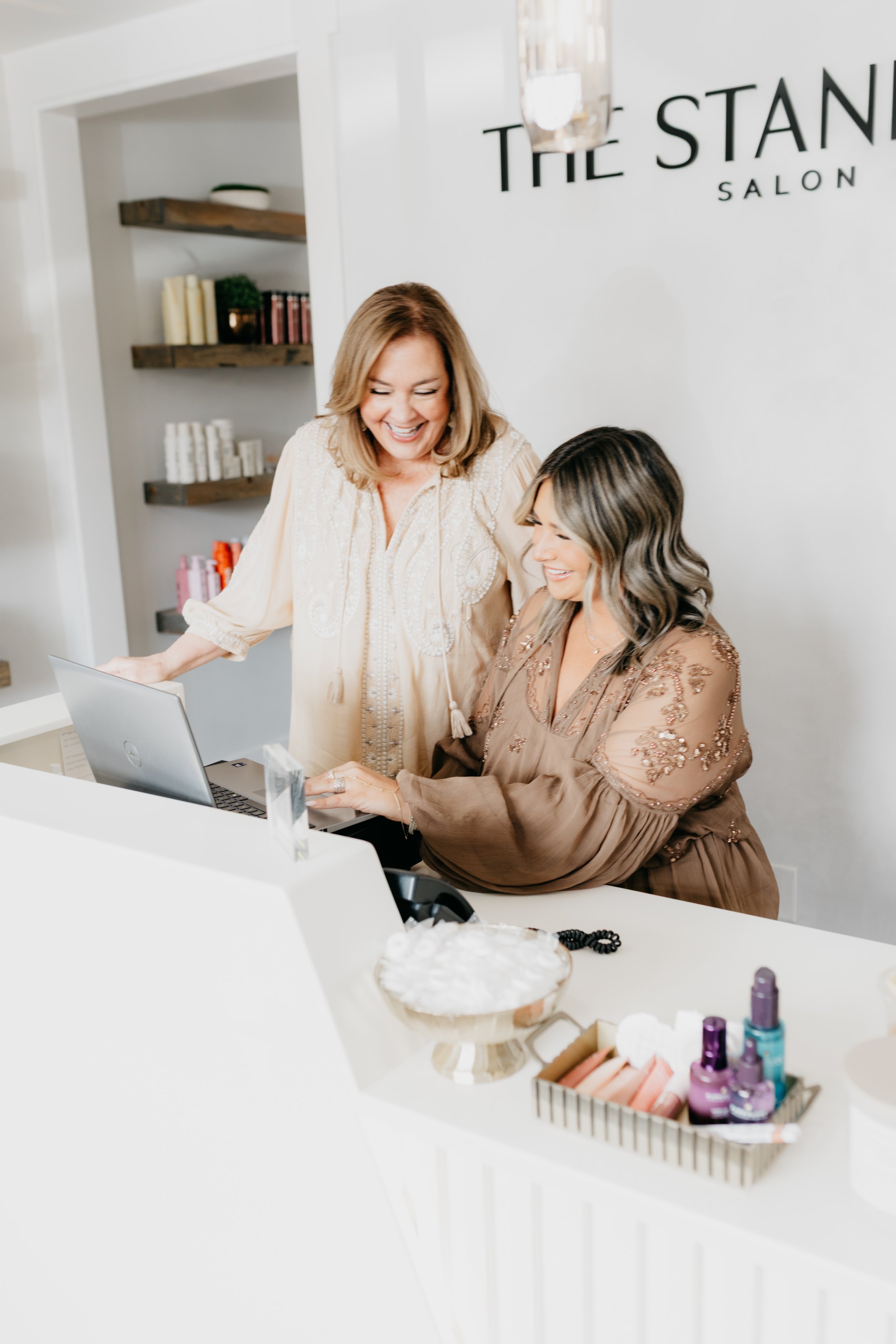 A woman in a brown dress and a woman in a beige blouse looking at a laptop, smiling, in a salon with shelves of beauty products in the background.