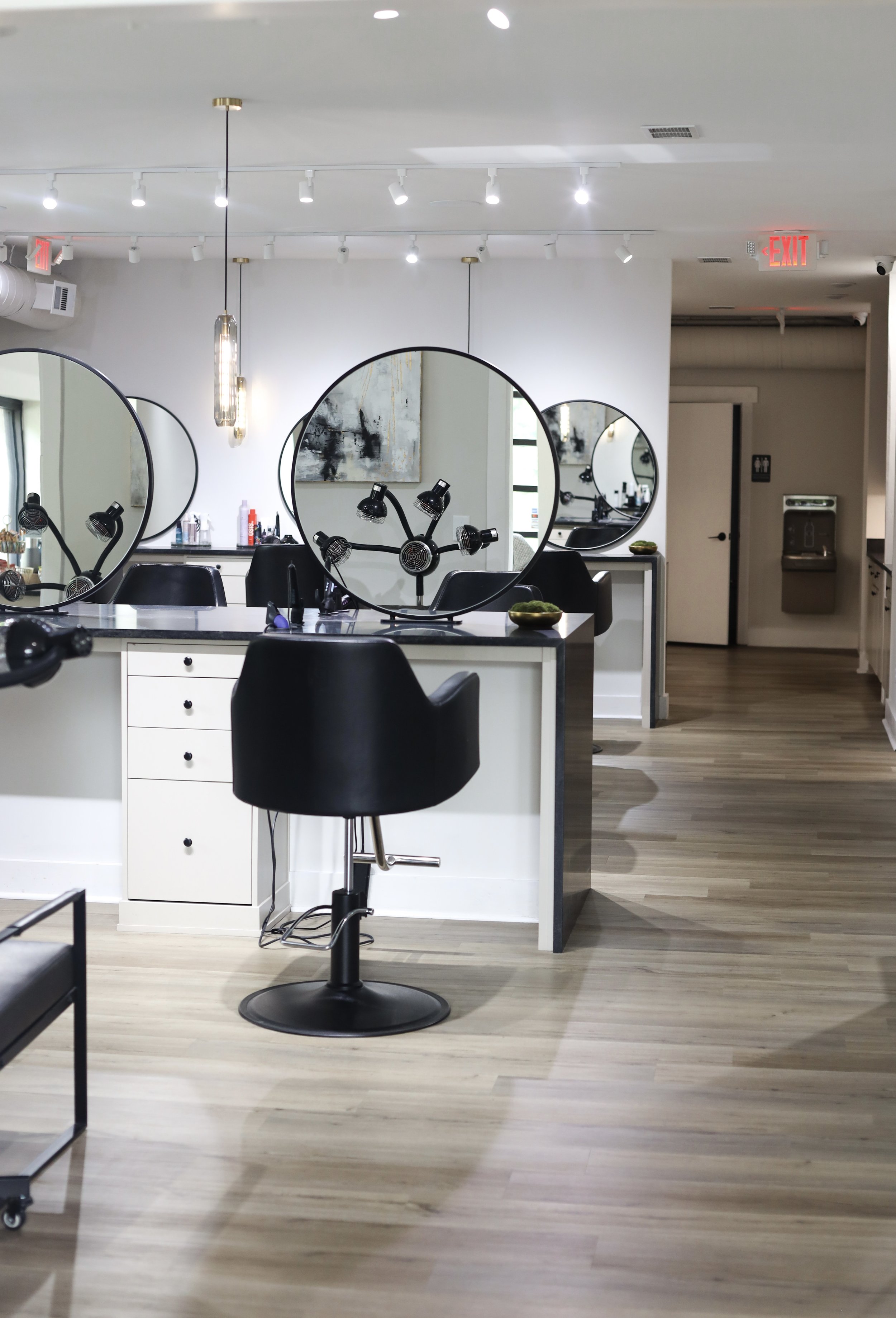 A modern hair salon interior with styling chairs, large round mirrors, and workstations with hair styling tools and products. The space features a white and black color scheme with wood flooring and contemporary lighting fixtures.