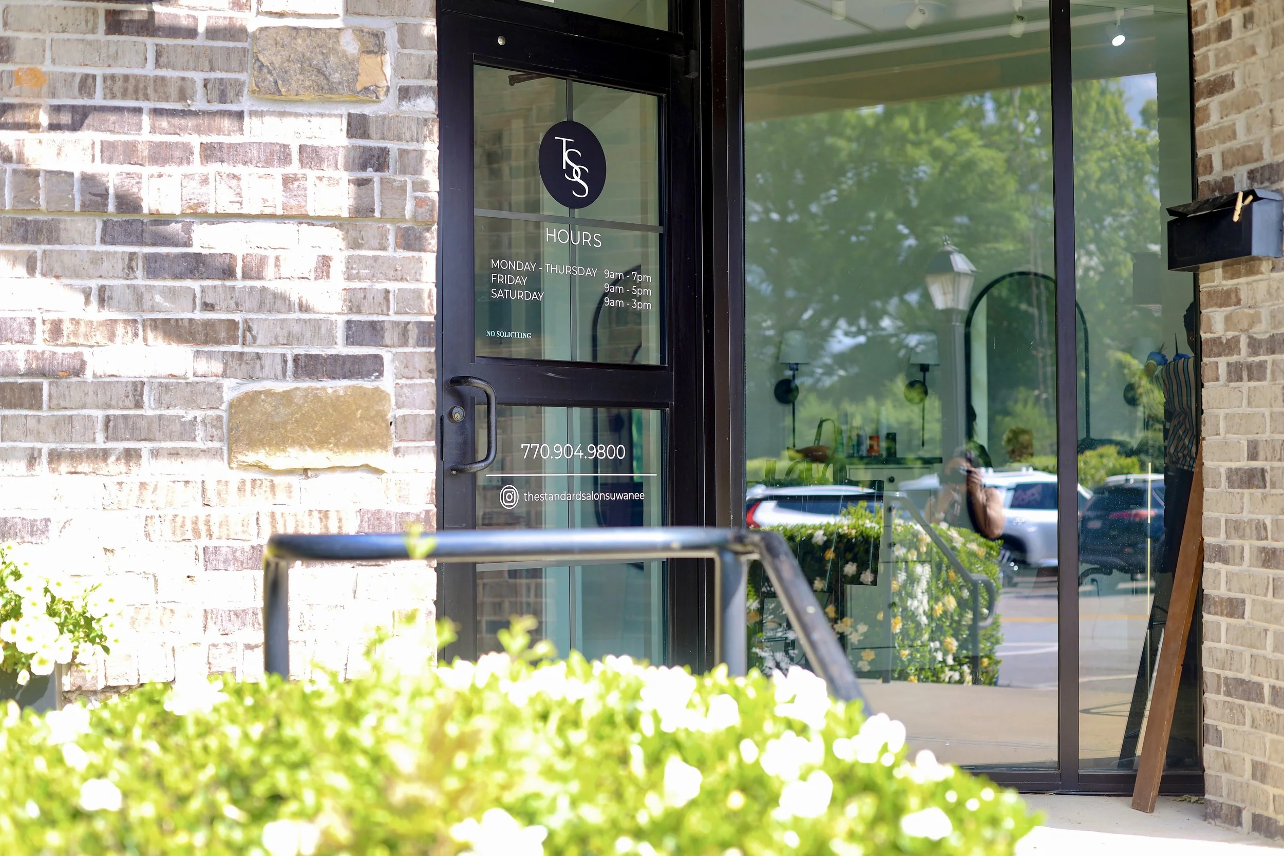 The entrance to a salon with a brick exterior, glass door, and window. The door has hours and contact information written on it, including social media icons. Inside, there are hanging lights, and a person can be seen taking a photo with a phone reflected in the window.