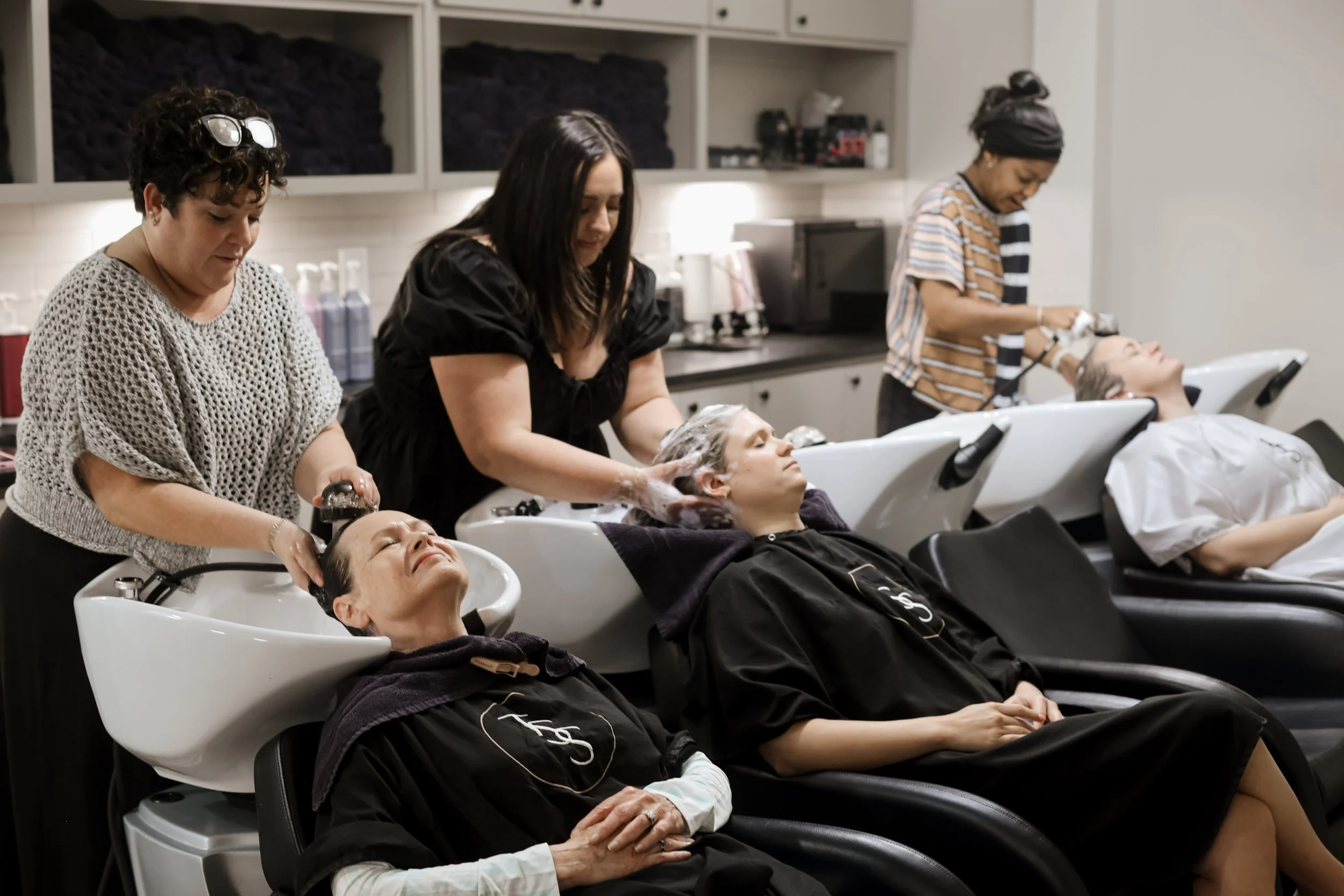 Three women are washing the hair of clients reclining in salon sinks in a hair salon.