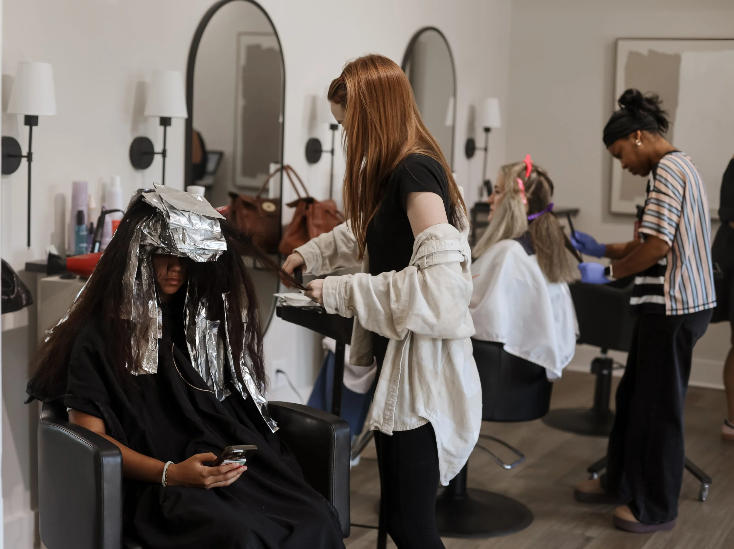 A woman with long dark hair covered in aluminum foil is sitting in a salon chair, looking at her phone. Behind her, a hairstylist with long, reddish hair is inspecting her hair. In the background, another hairstylist with black hair tied up, is working on another client's hair, with some clients having colored hair or accessories.