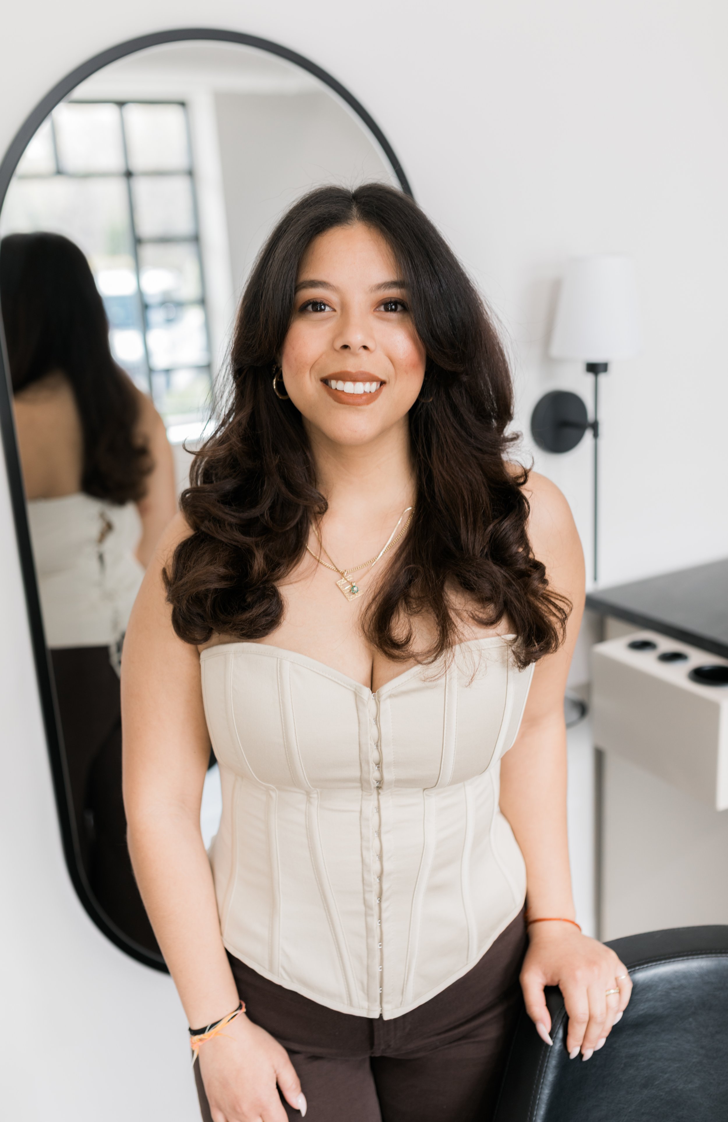 A woman with long dark hair smiling in a plaid blue and white dress with a yellow off-white Off-White belt, standing indoors near a white wall.