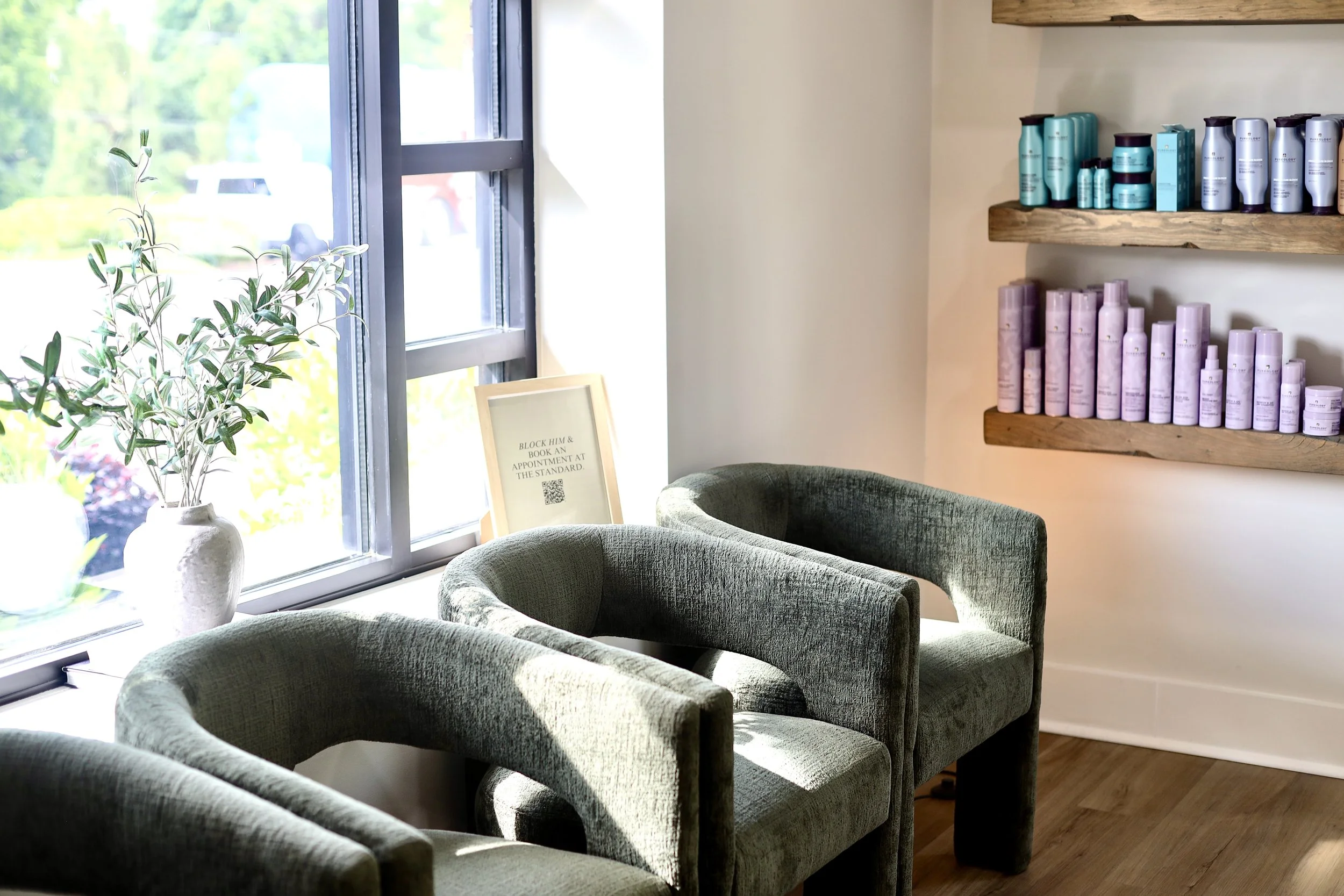 A cozy seating area with curved green chairs near large windows, wooden shelves with hair care products on a beige wall, and a framed sign on a table.