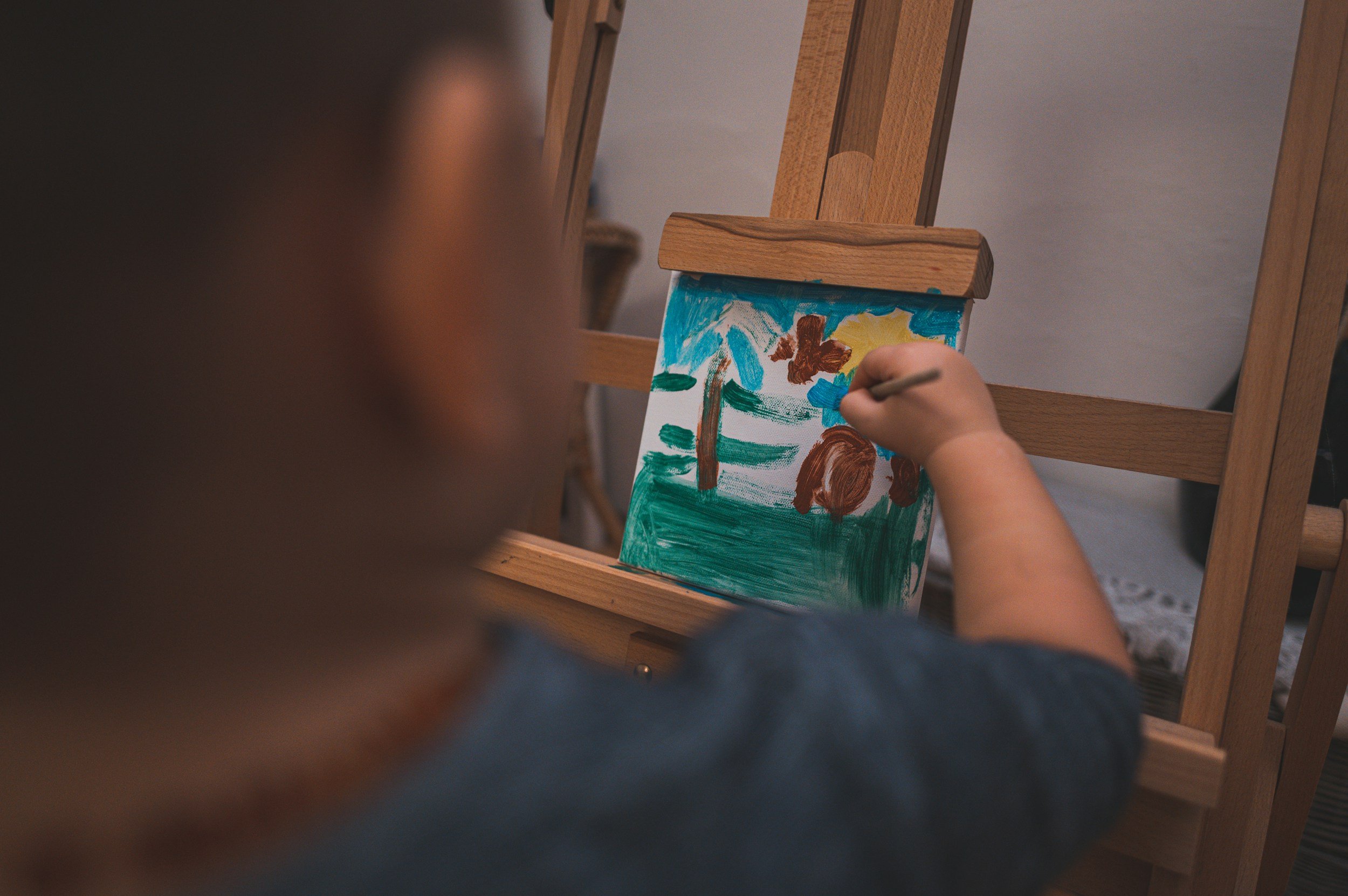 Child painting a colorful picture of a tree, sun, clouds, and a butterfly on a canvas.