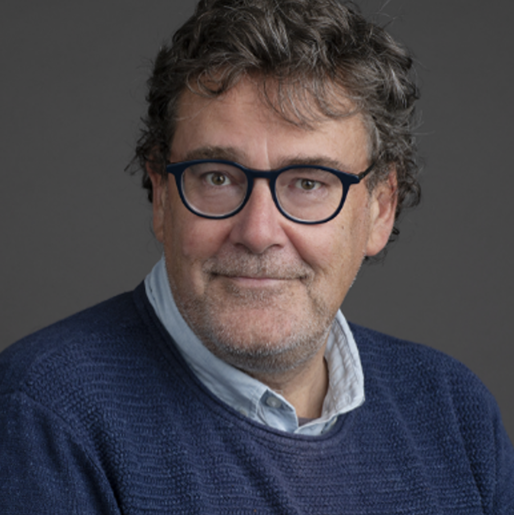 Portrait of a middle-aged man with curly brown hair, glasses, a white collared shirt, and a navy blue sweater against a dark gray background.