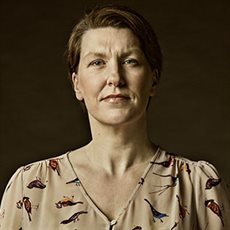 Portrait of a woman with short dark hair, wearing a light-colored blouse with a bird pattern, set against a dark background.