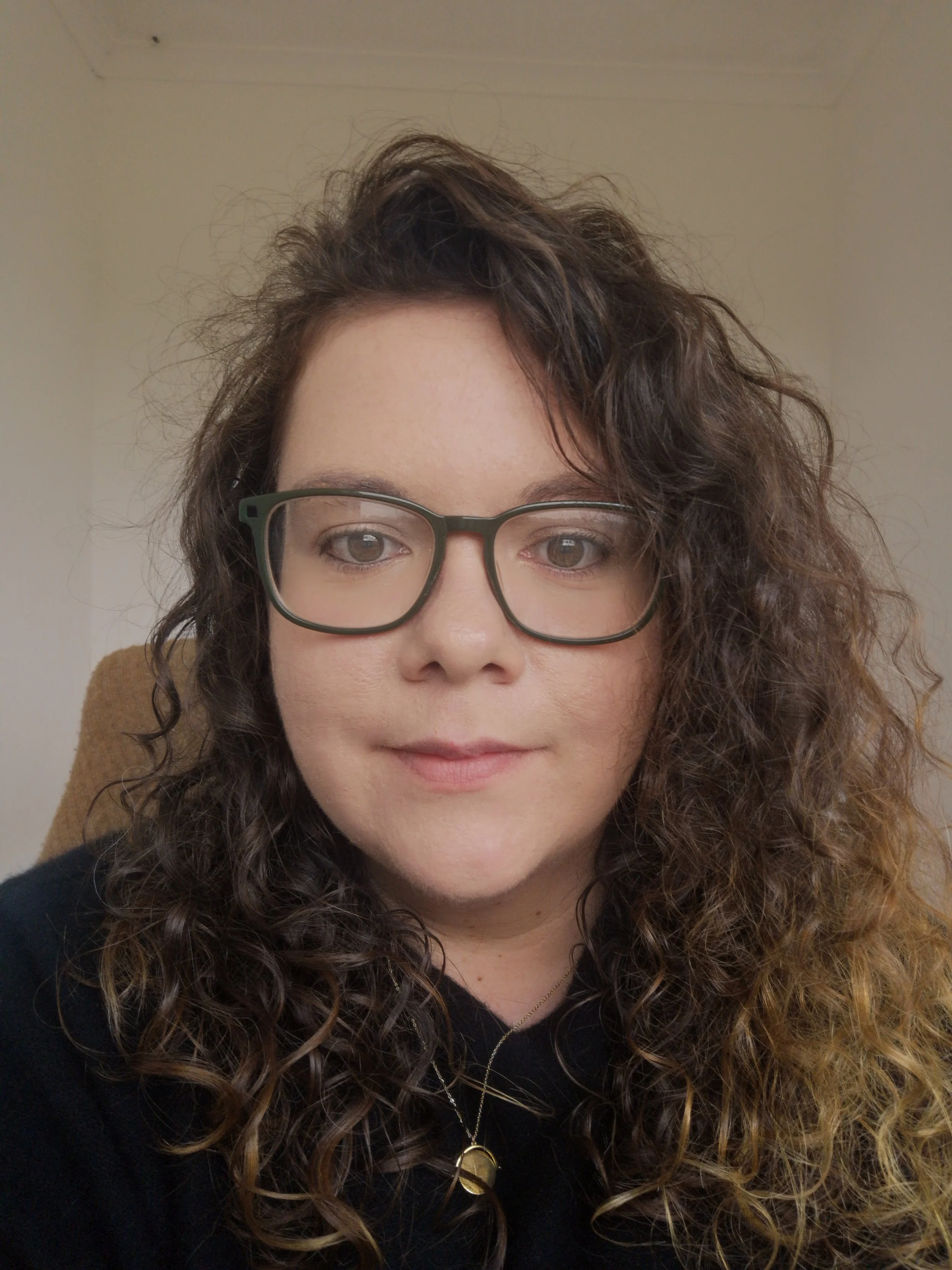 Close-up of a woman with curly brown hair, wearing glasses with dark green frames, a black top, a gold necklace, and a gold pendant, looking into the camera. The background is plain.